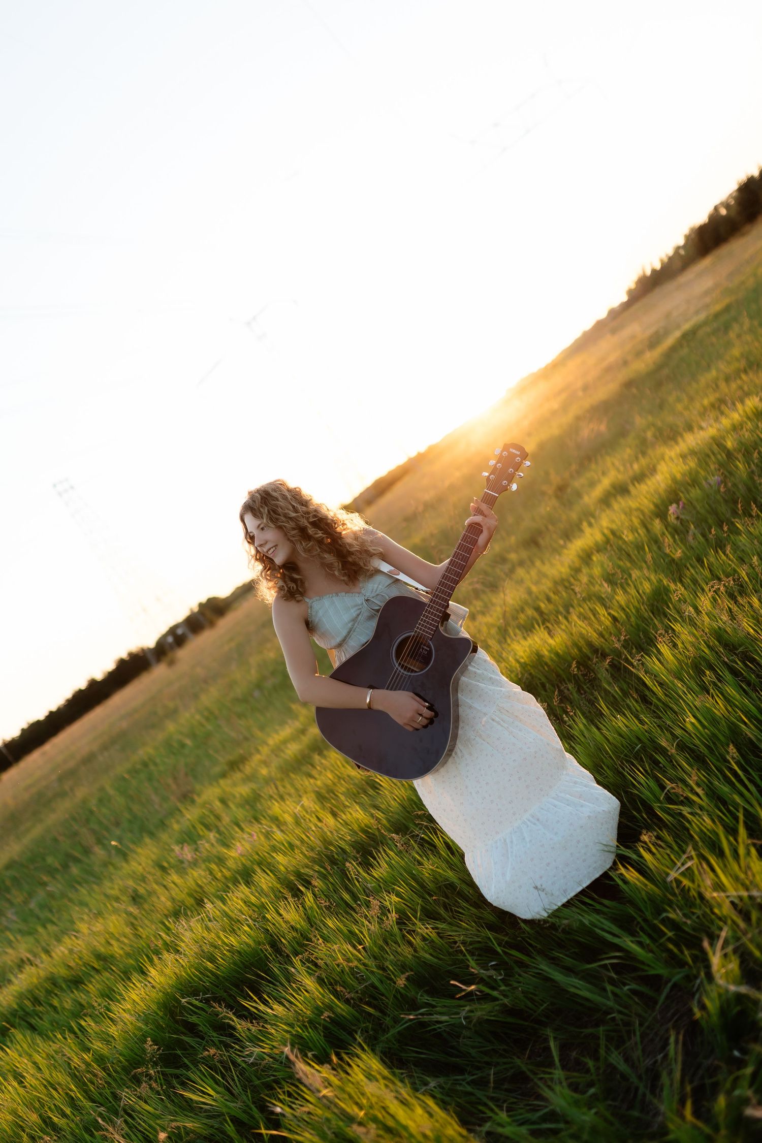 Curly-haired woman in white dress holding acoustic guitar in a sunlit green meadow at sunset.