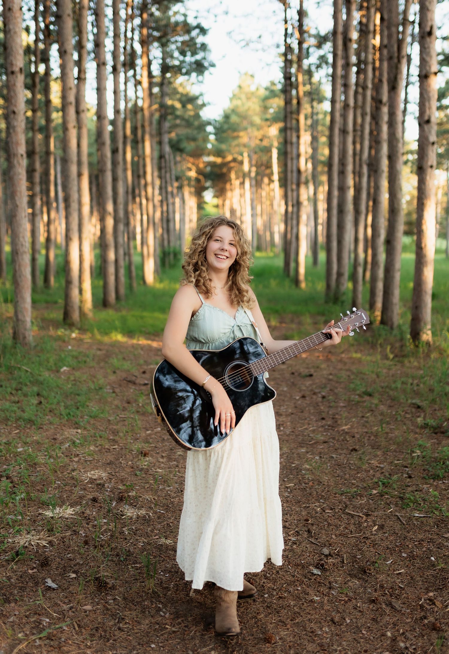 Happy young woman in mint top and white skirt playing guitar while walking through a pine forest.