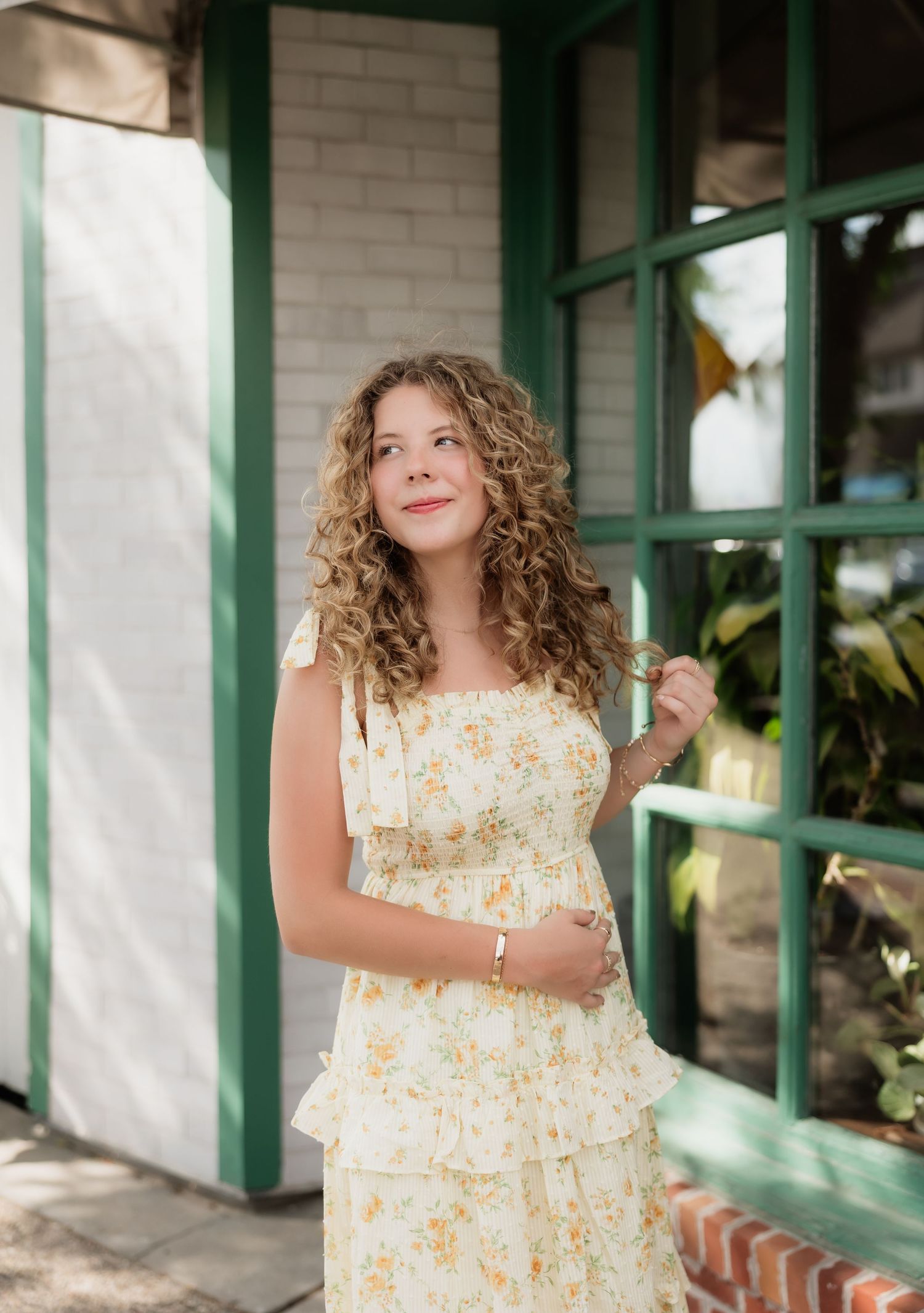Young woman in a yellow floral tiered sundress gazing upward beside a green-trimmed window.