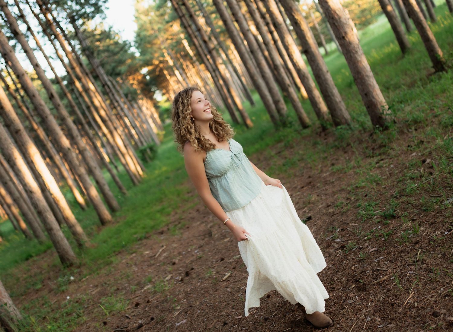 Young woman in light blue and white dress walking through a pine forest in golden sunlight.
