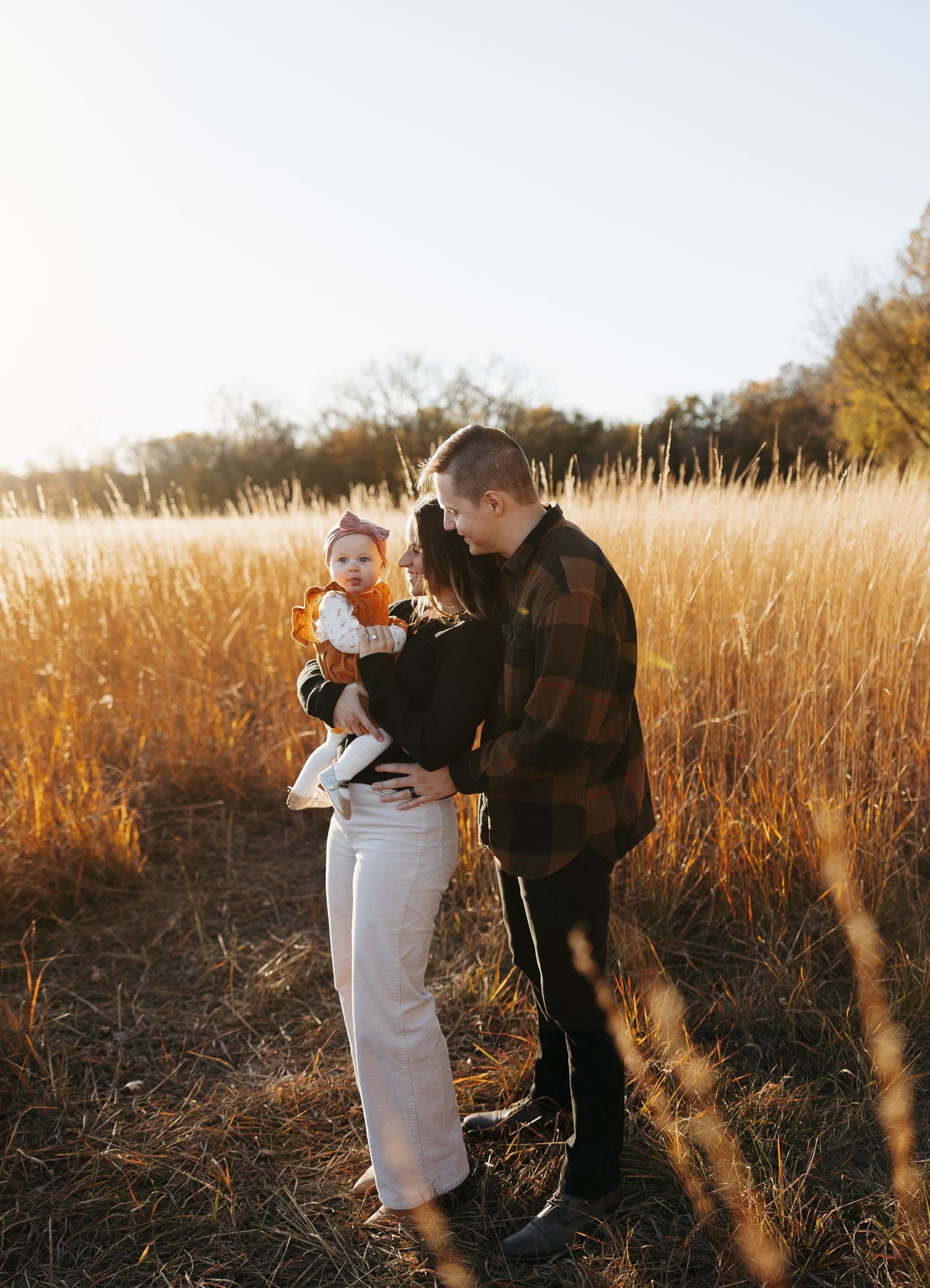 Young couple holding baby girl in orange dress standing in golden autumn meadow at sunset.