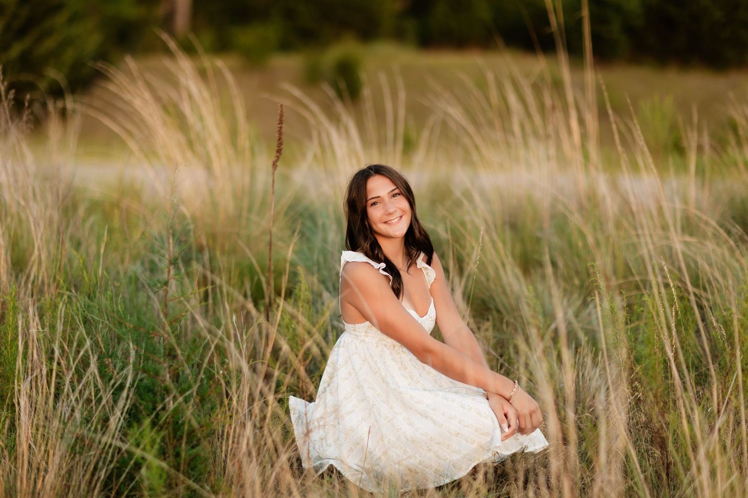 Brunette girl in white dress crouching and smiling among tall golden grasses.