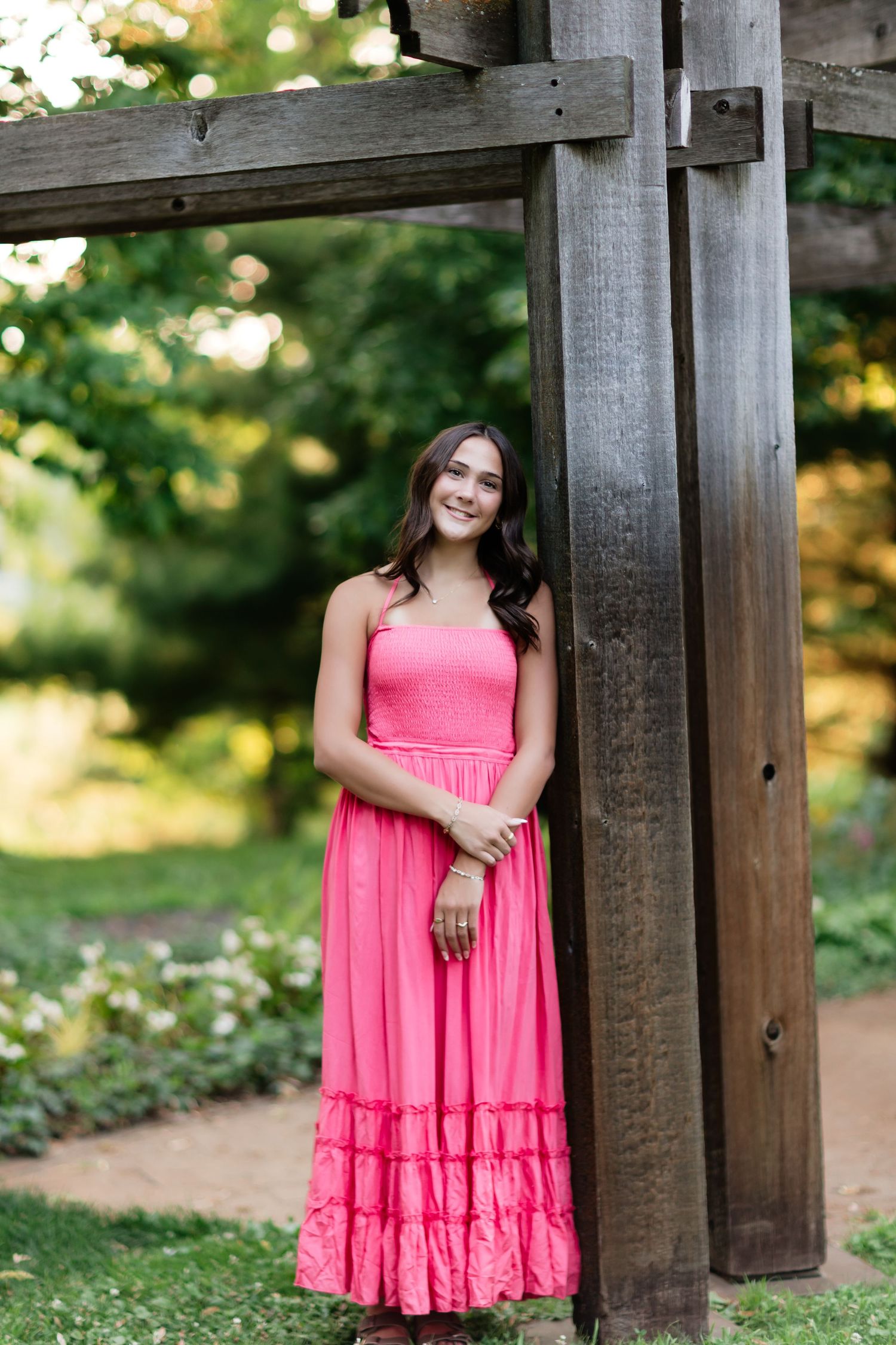 Young woman in pink maxi dress smiling outdoors near wooden pergola in lush green garden.