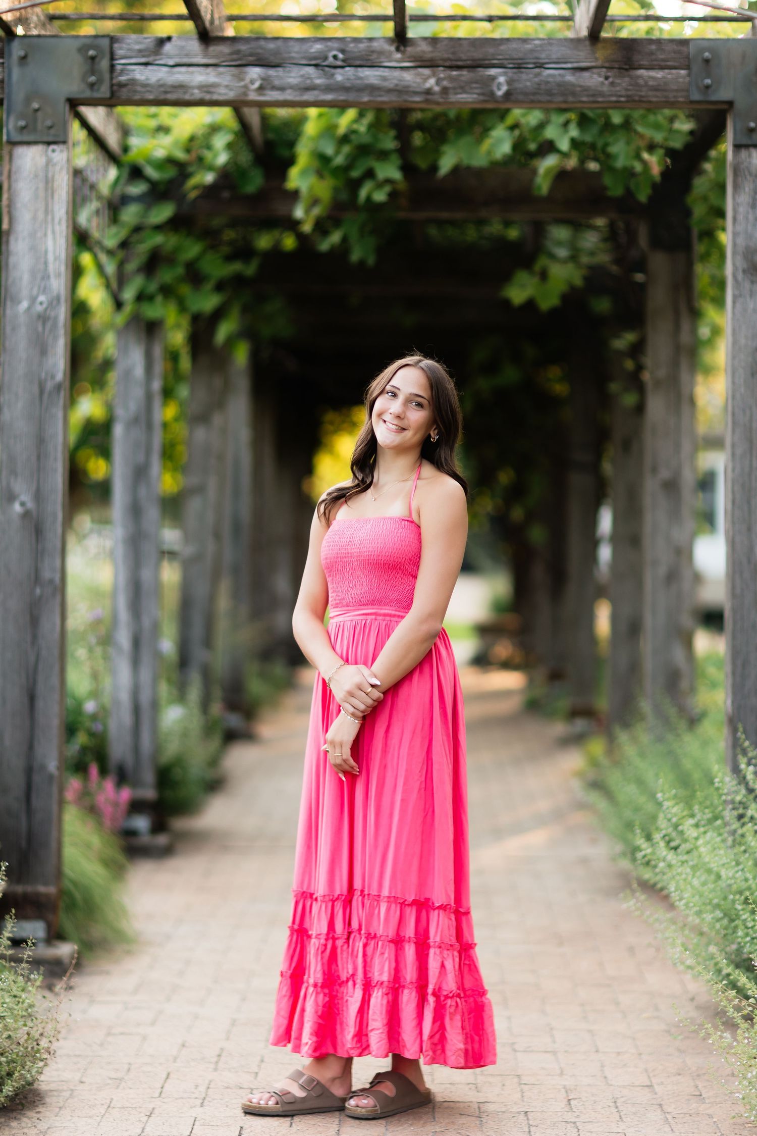 Brunette teen in hot pink maxi dress standing under wooden pergola draped with green vines.