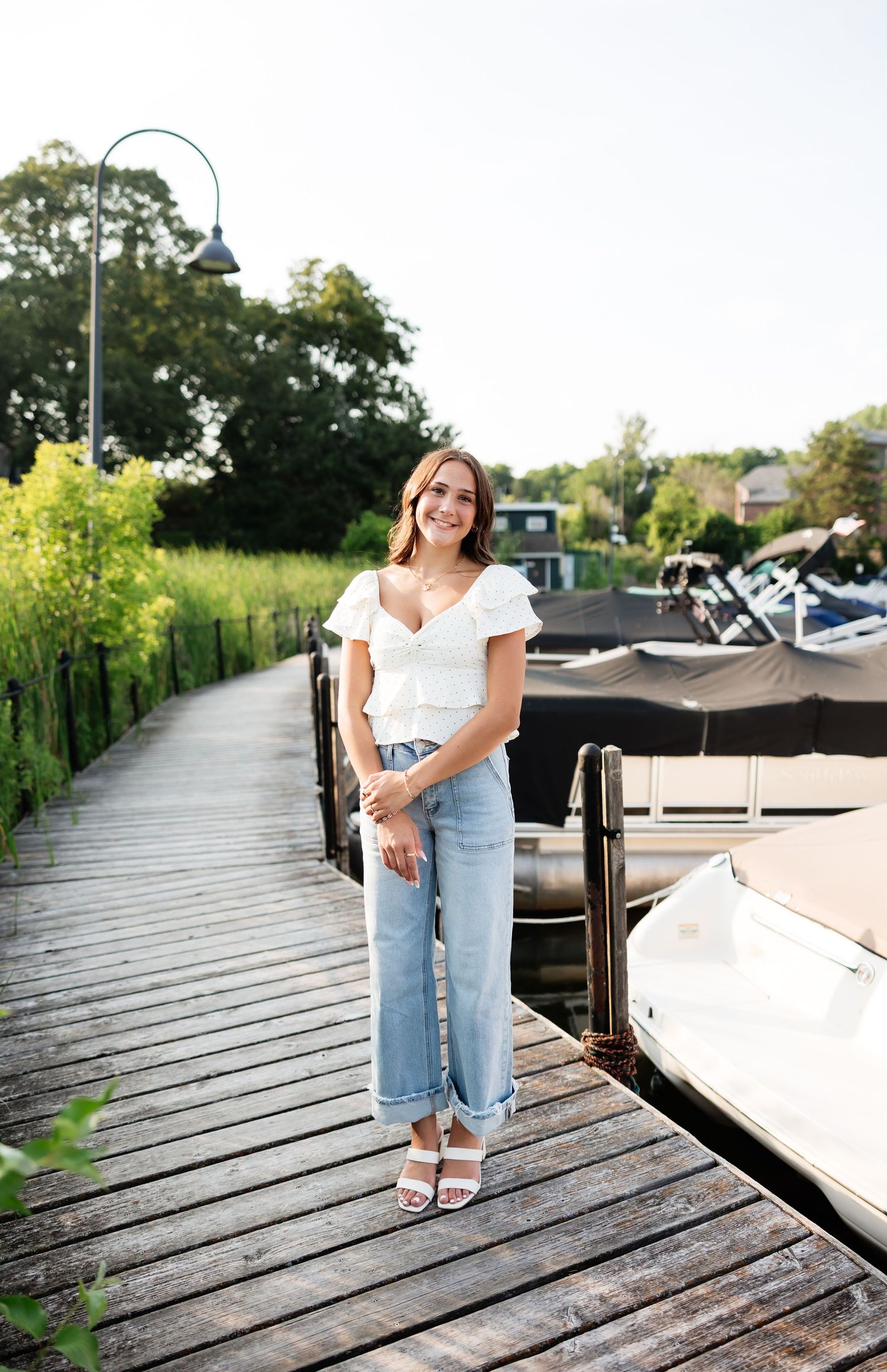 Young woman in white top and light jeans smiling on wooden dock with boats and greenery behind her.