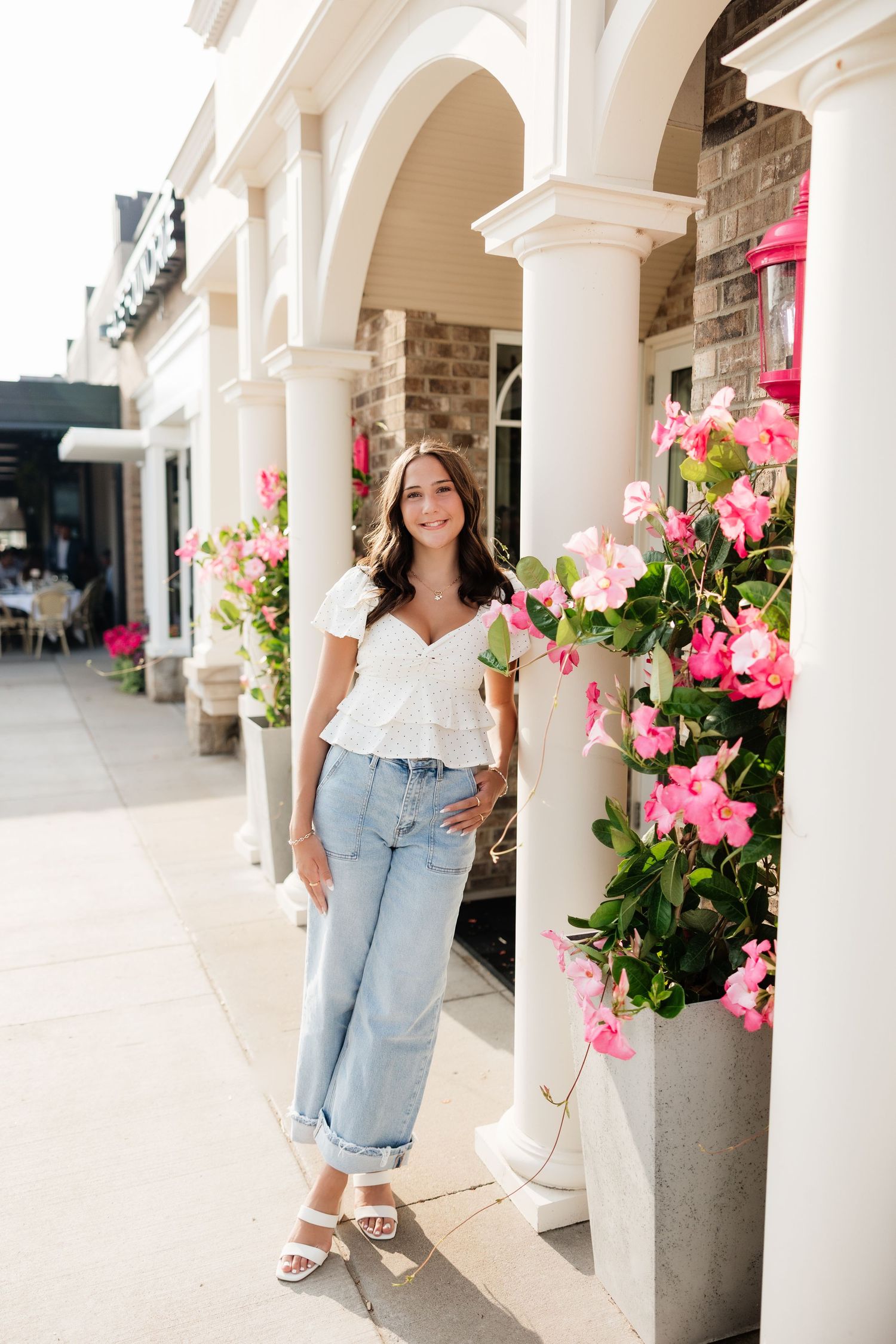 Smiling brunette woman in white top and jeans posing by pink flowers and white columns.