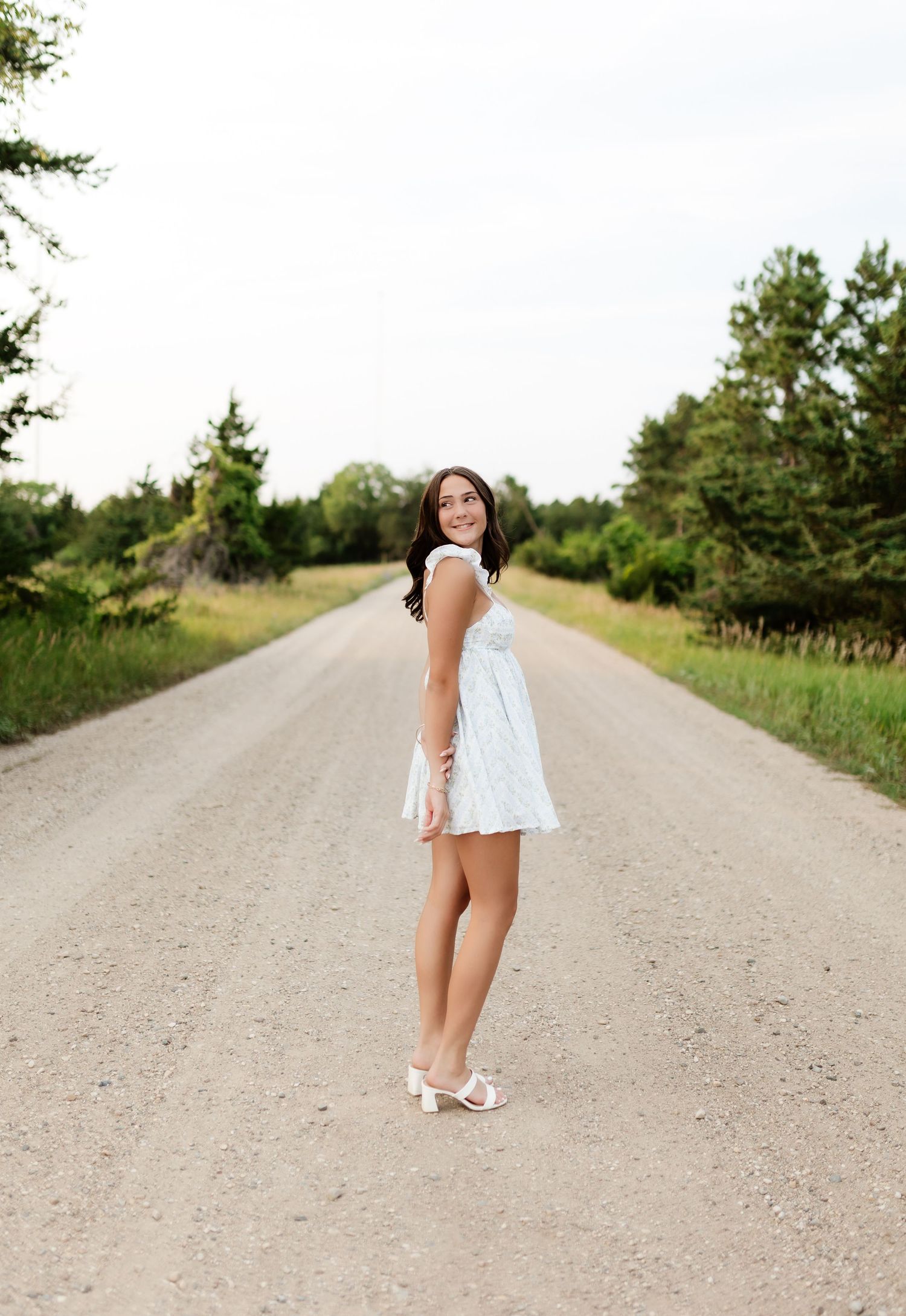 Smiling young woman in a white dress stands on a peaceful gravel road surrounded by lush green trees.