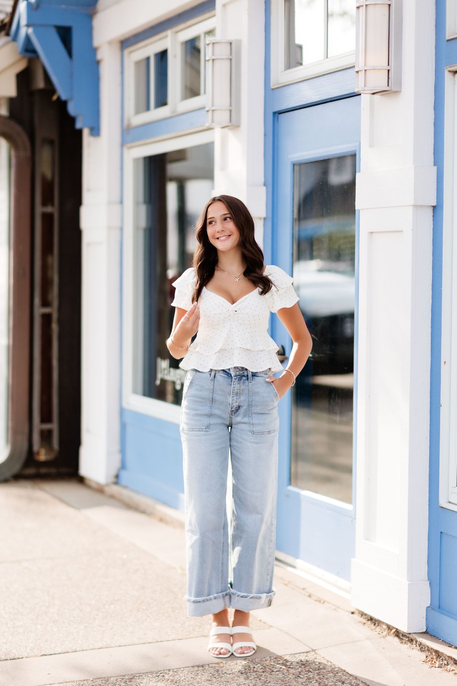 Young woman in white ruffle top and cuffed wide-leg jeans poses confidently outside blue and white building.