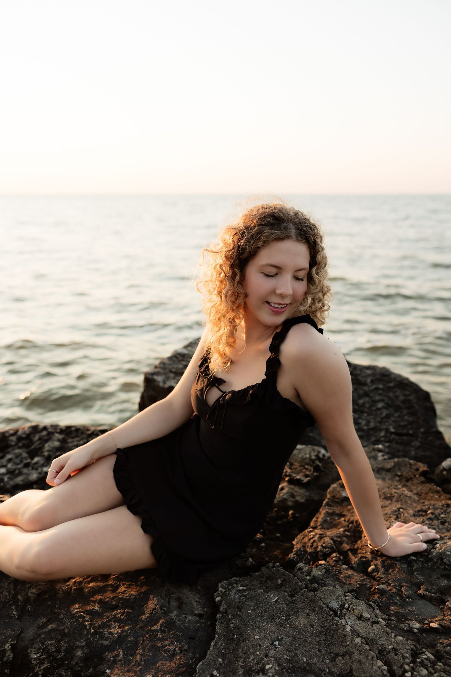 Young curly-haired woman in black dress sitting on rocks by the ocean at golden hour sunset.