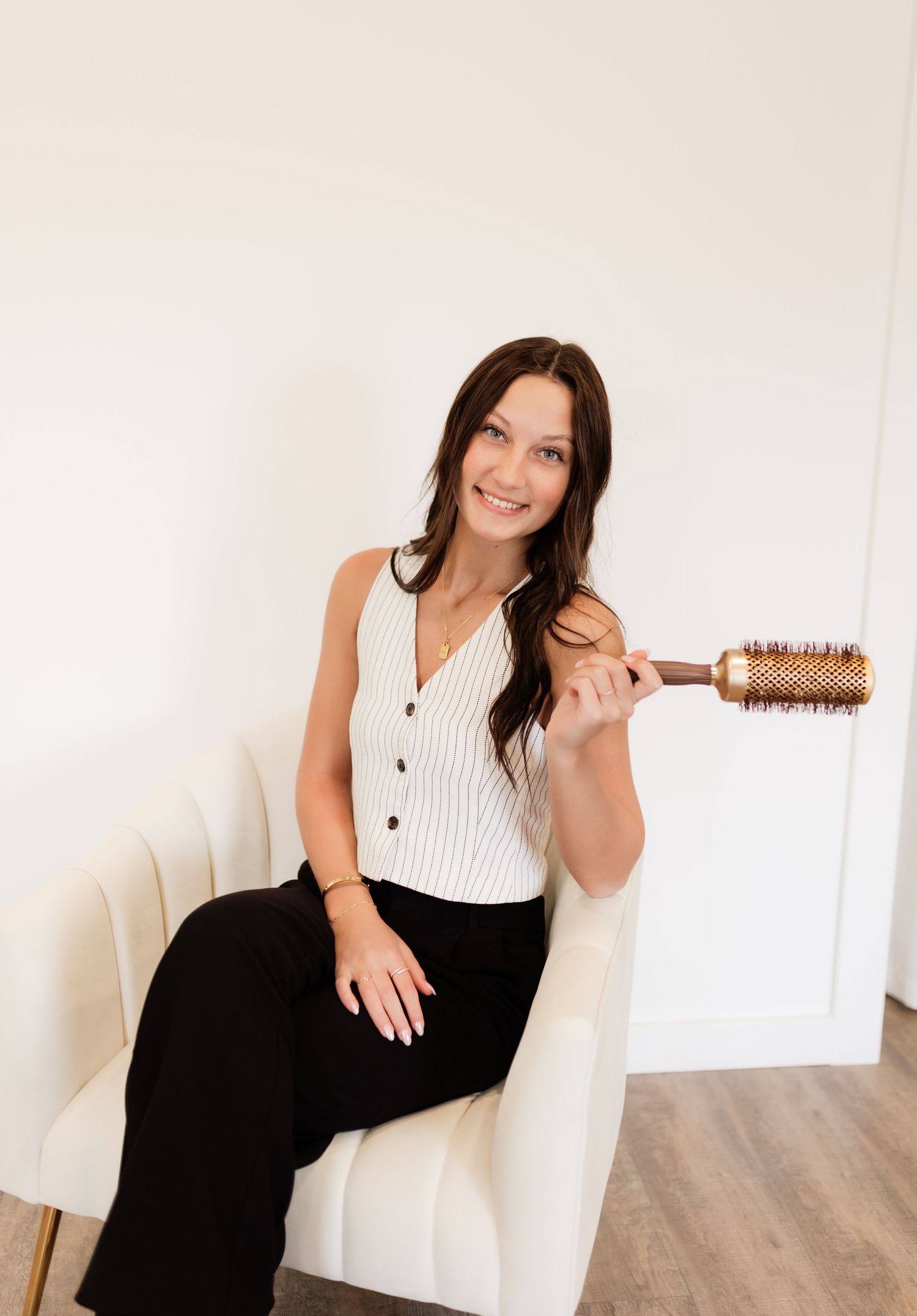 Smiling brunette woman sitting in cream chair holding a round barrel hair brush in a bright studio setting.