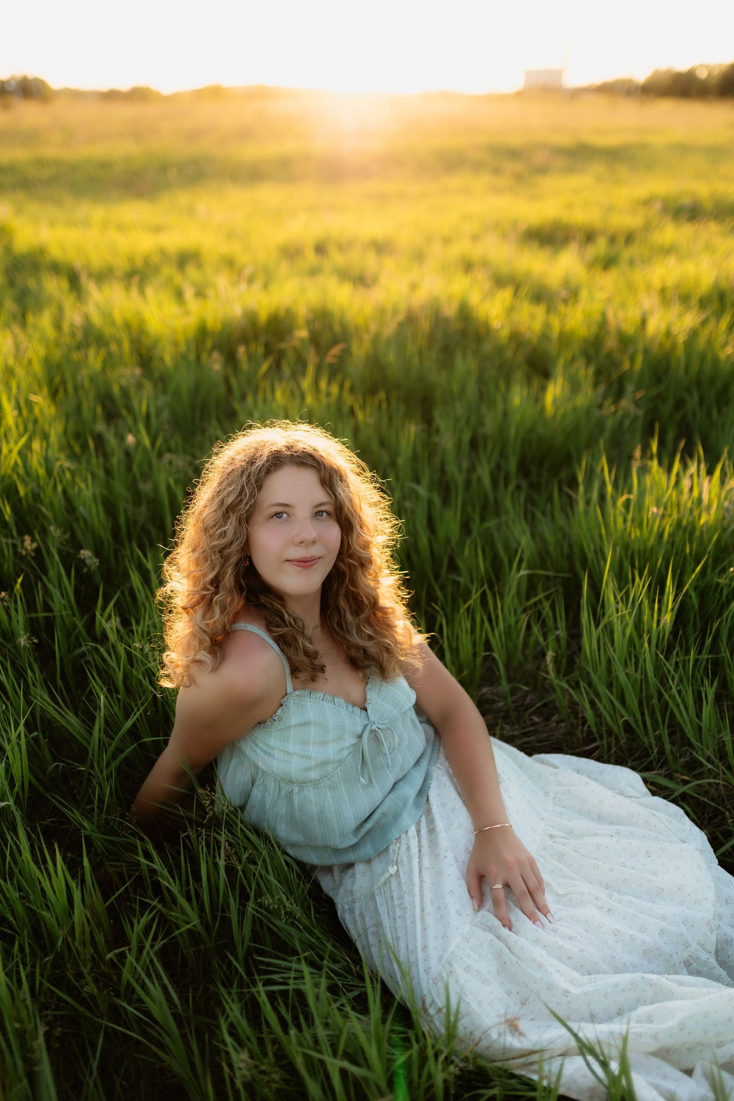 Young woman with curly hair in a light blue dress sitting in a green field during golden hour sunset.