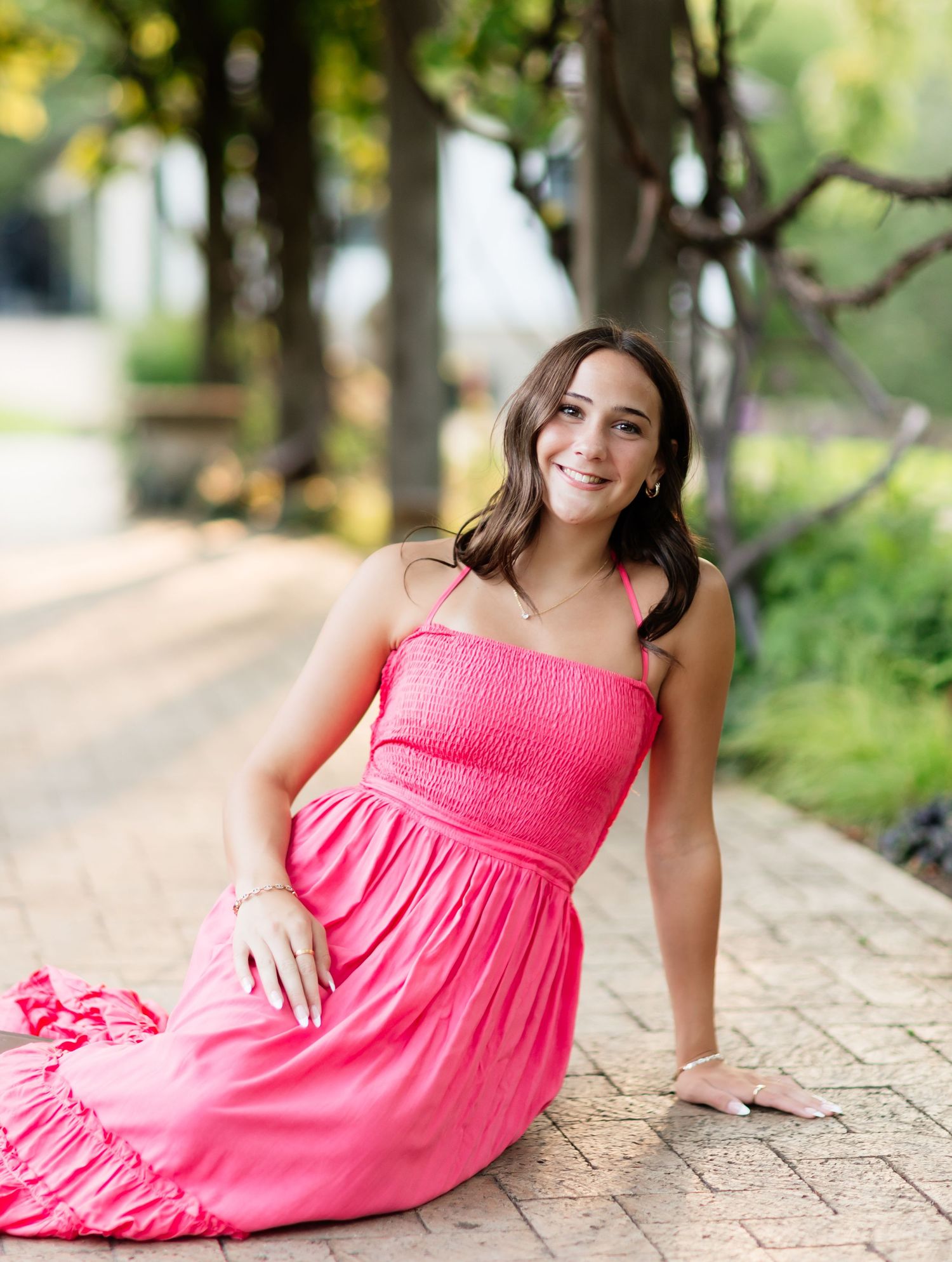 Smiling young woman in bright pink smocked maxi dress sitting on a brick pathway in a park.
