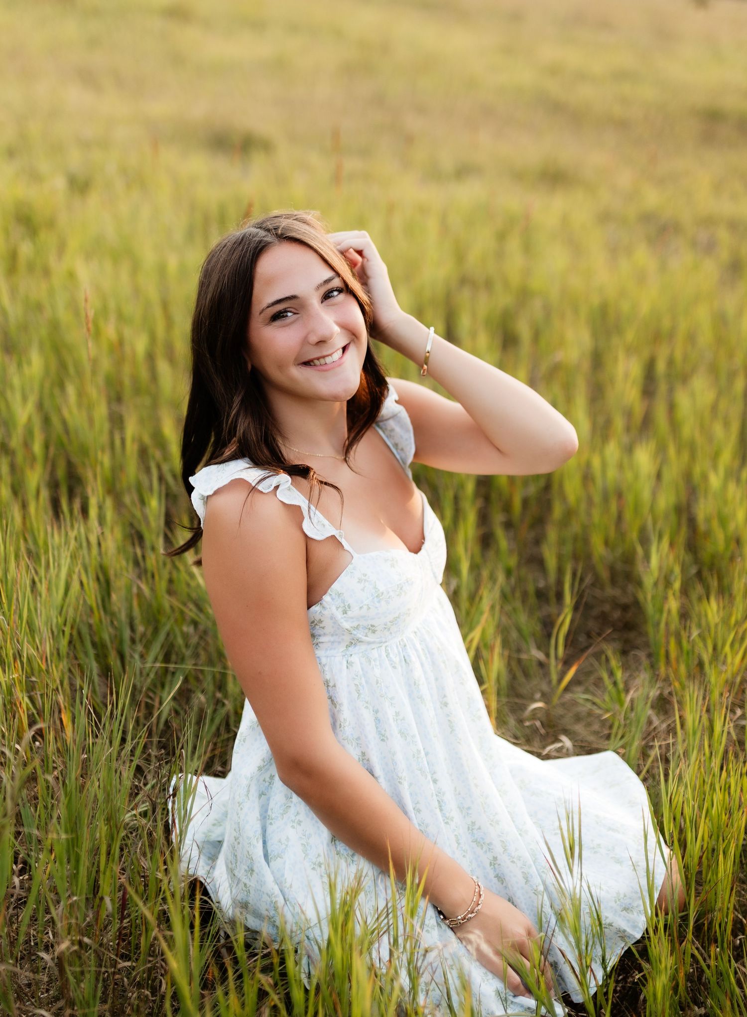 Smiling young woman in light blue dress sitting in tall green grass field outdoors.