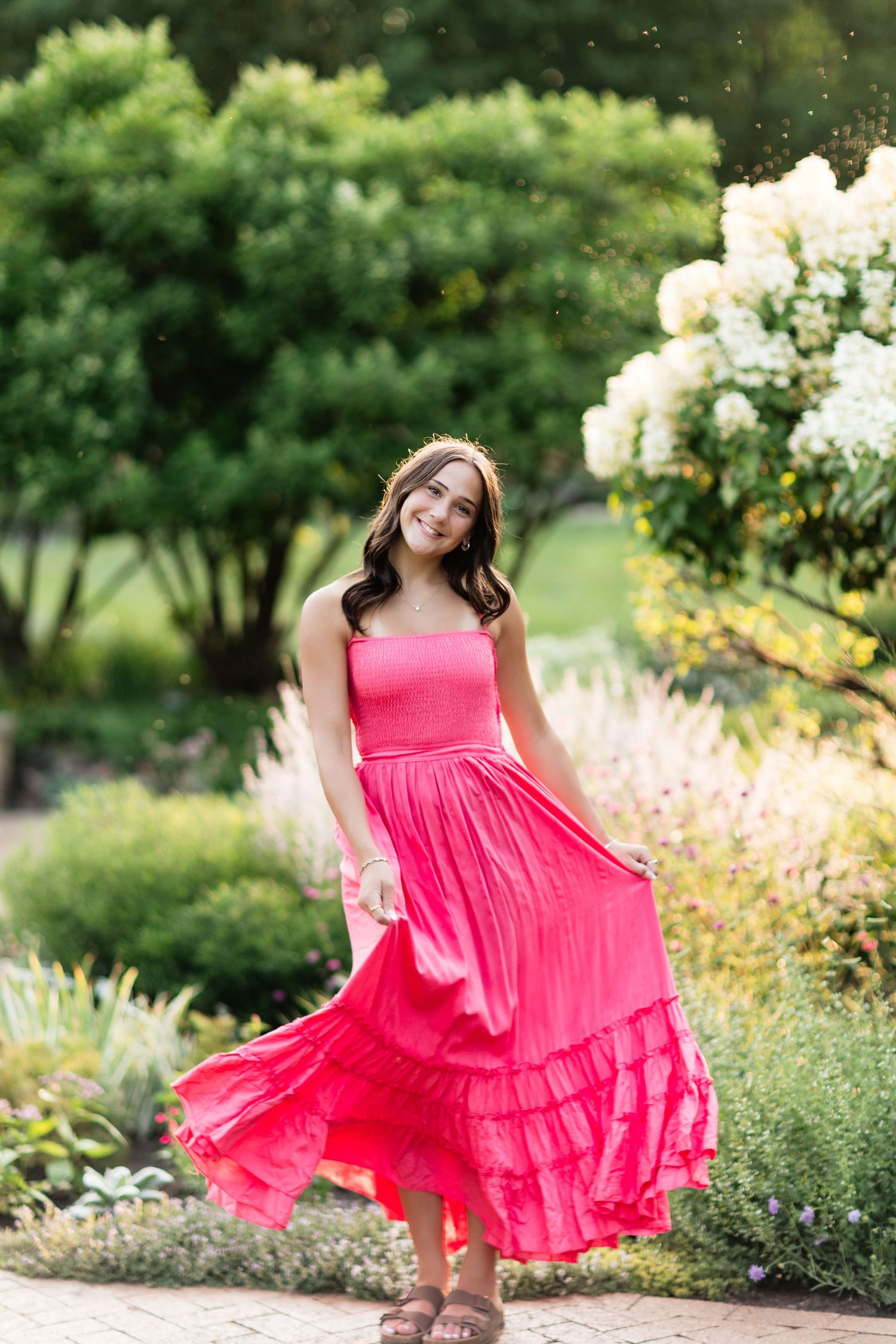 Smiling young woman twirling in a bright pink strapless maxi dress in a lush garden setting.