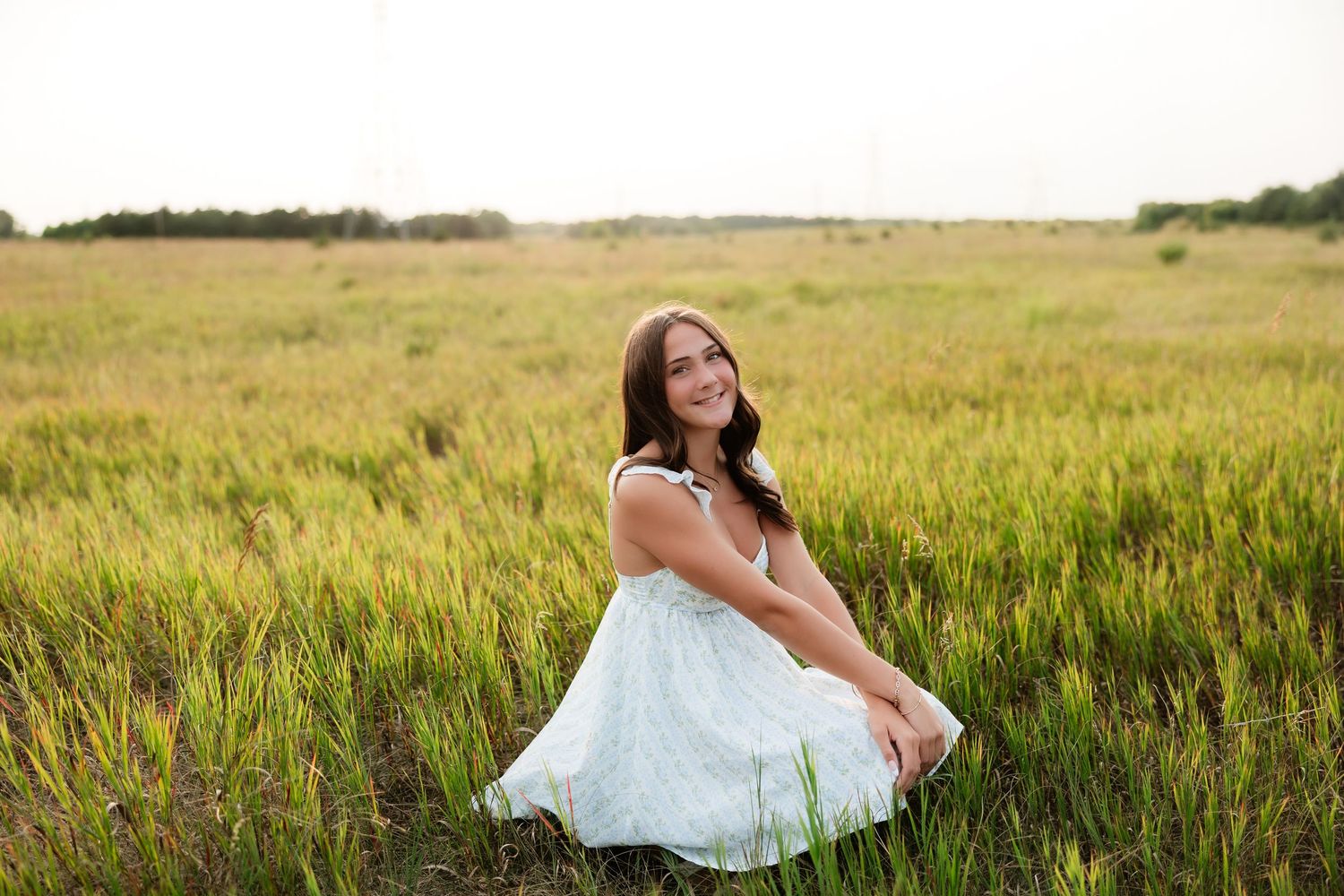 Young woman in white sundress sitting in lush green field under soft overcast sky.