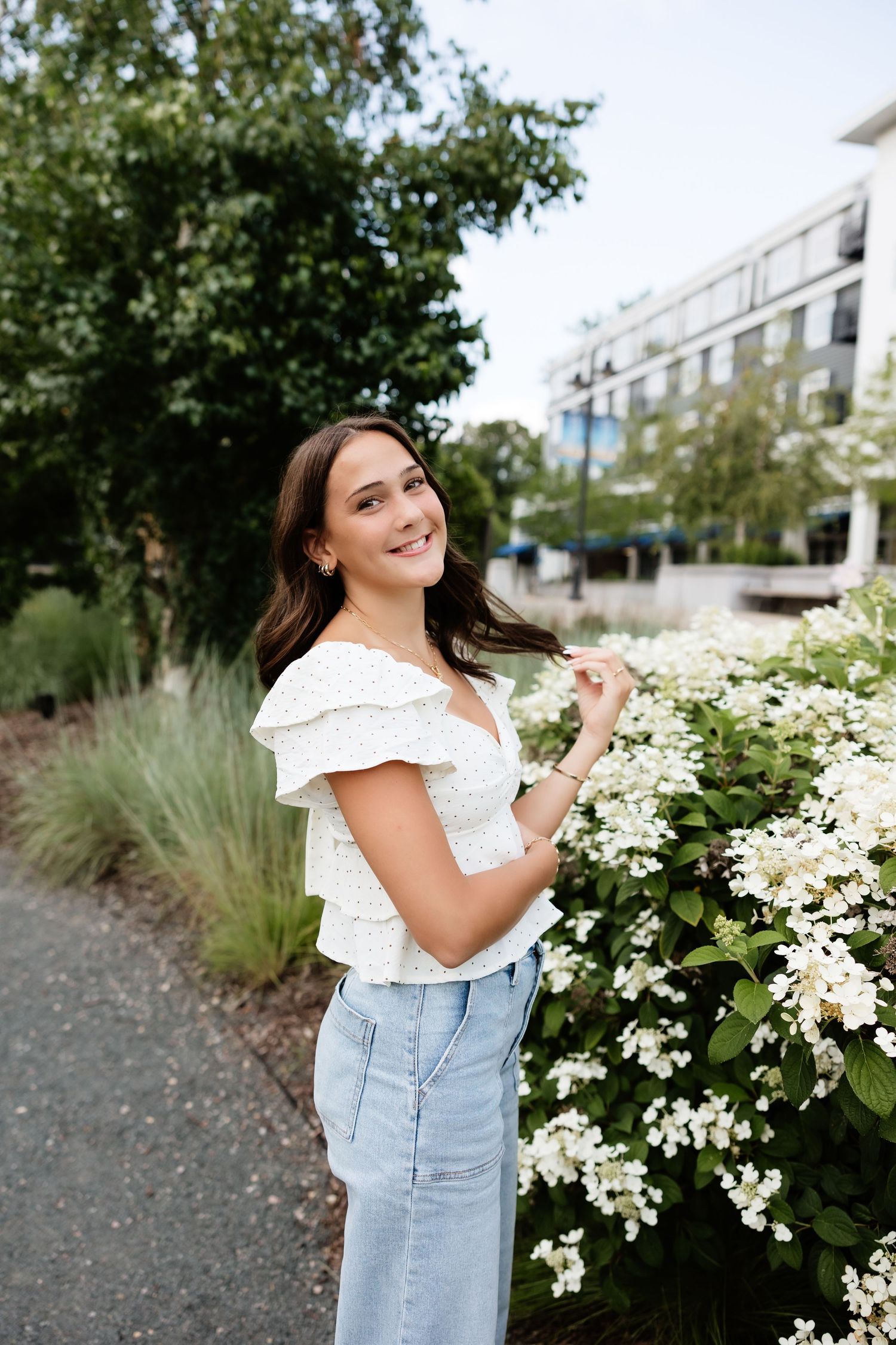 Young woman in white ruffled top and denim skirt smiling beside white hydrangea blooms outdoors.