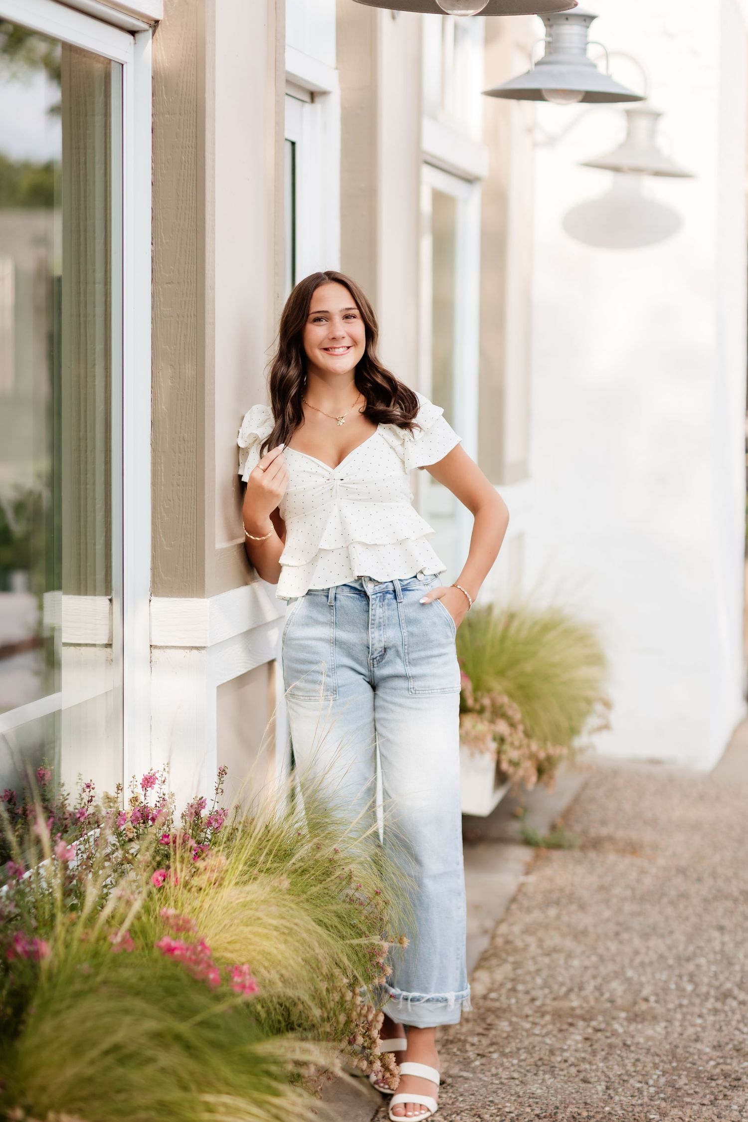 Smiling woman in white top and wide-leg jeans standing outside shop with hanging lantern and flowers.