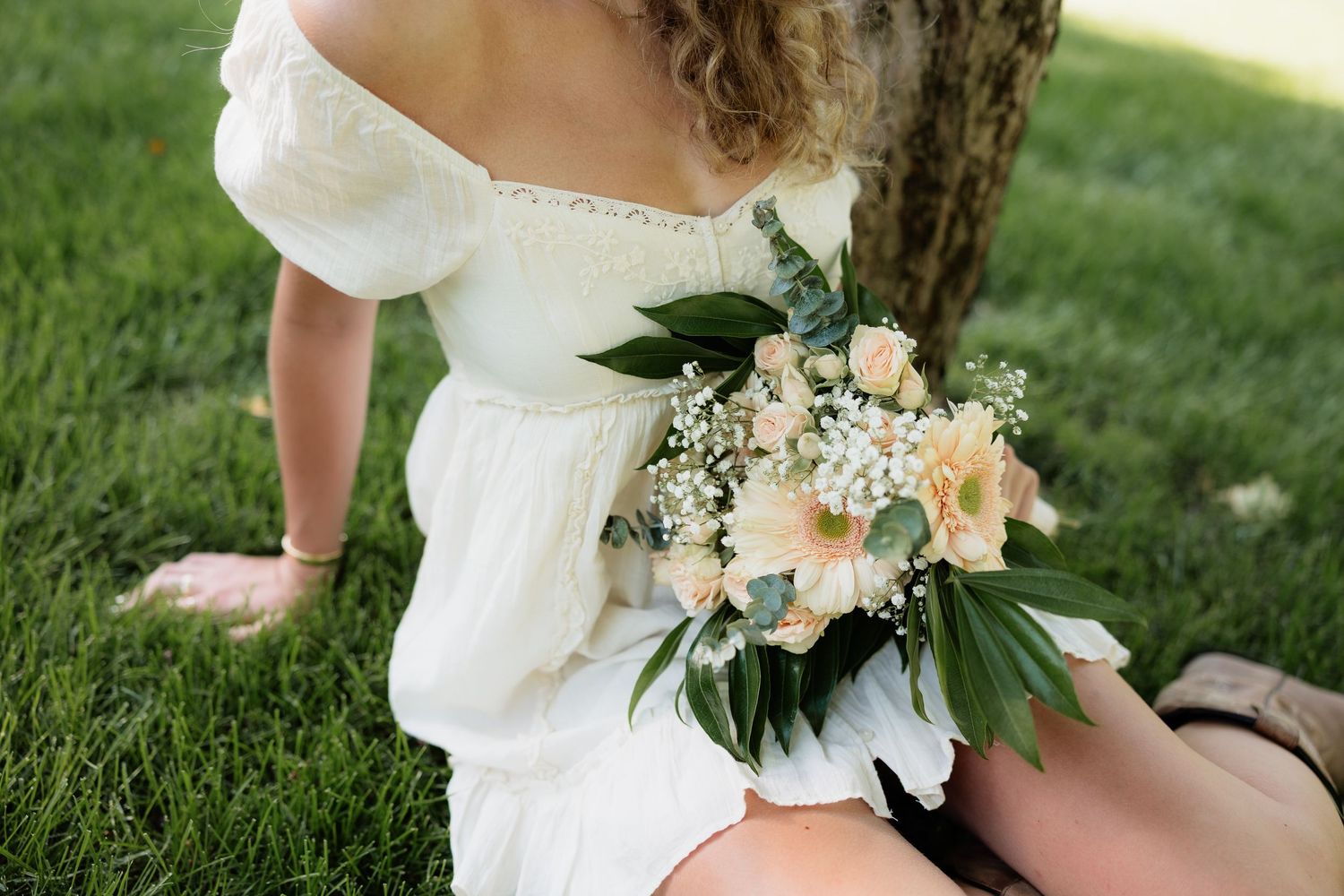 Woman in white off-shoulder dress holding a delicate bouquet of peach roses and greenery while sitting on grass.