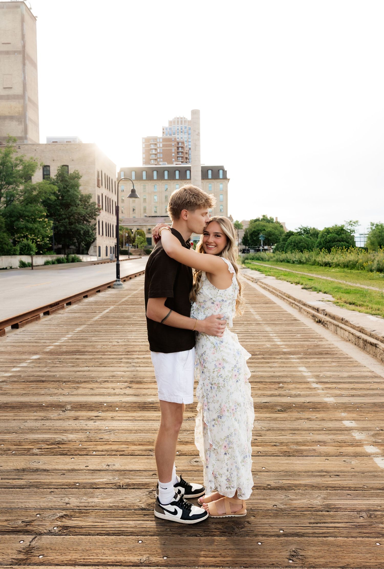 A couple hugging on a wooden railroad bridge, man in black shirt and white shorts, woman in floral maxi dress, city backdrop.