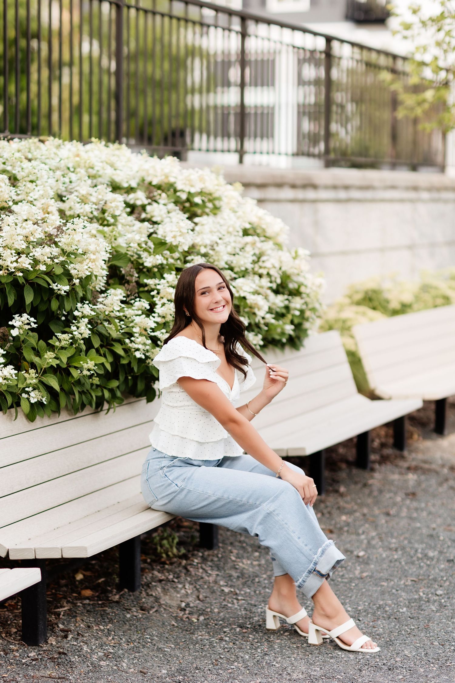 Young woman in white top and jeans sitting on bench surrounded by white hydrangeas, smiling outdoors.