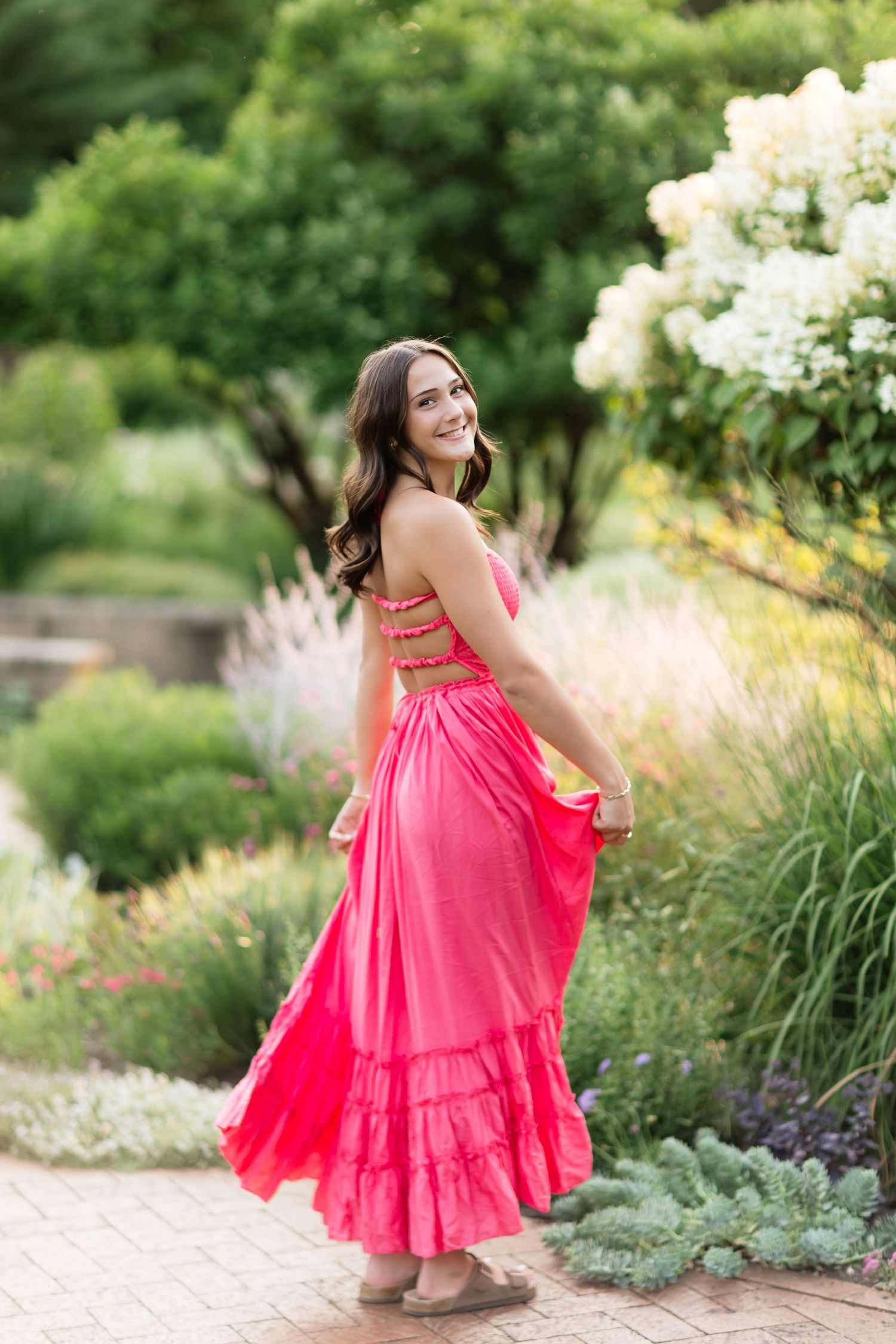 Young woman in pink maxi dress twirling in a lush garden with blooming flowers.