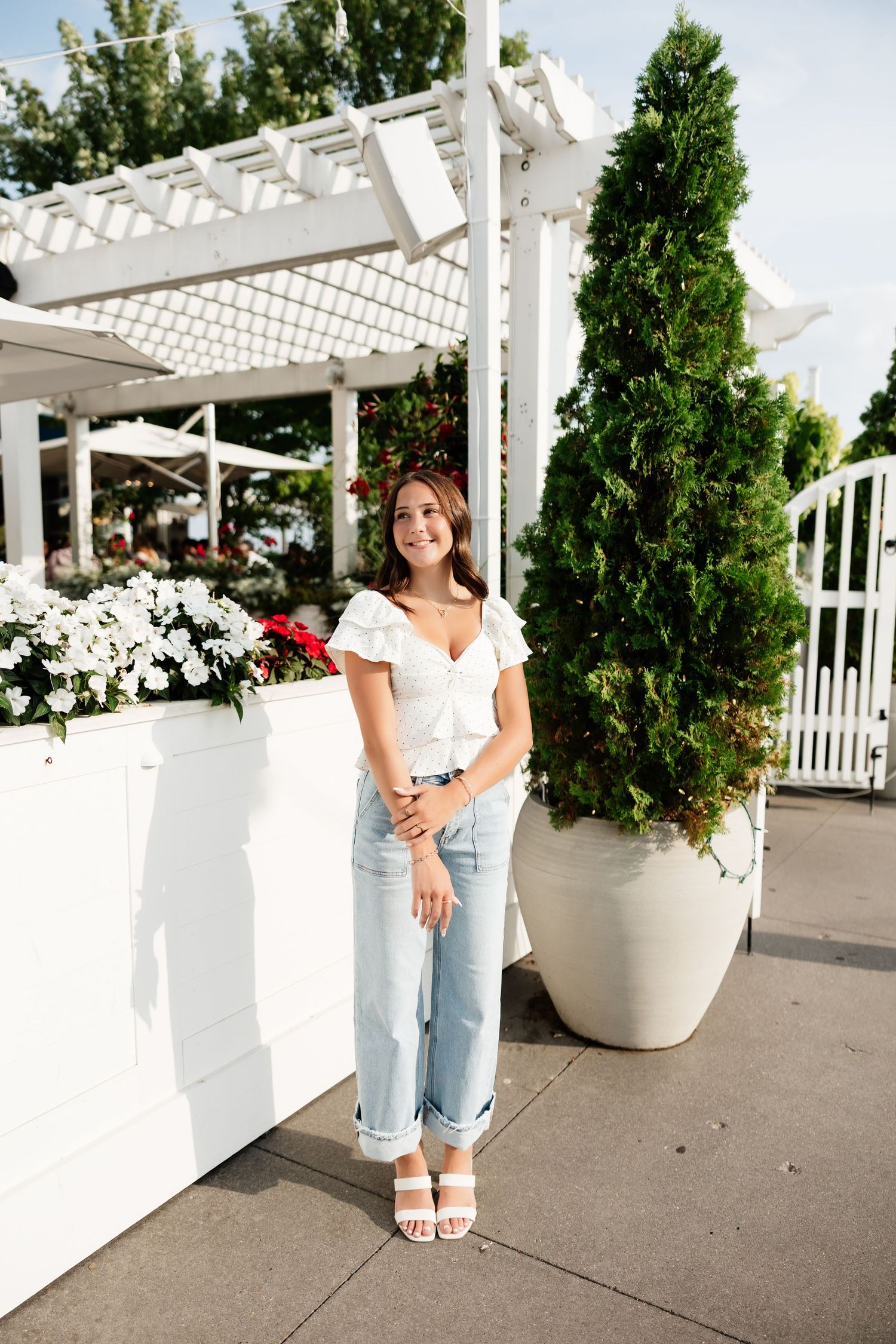 Young woman in white crop top and light jeans smiling outdoors near white planter boxes and evergreen tree.