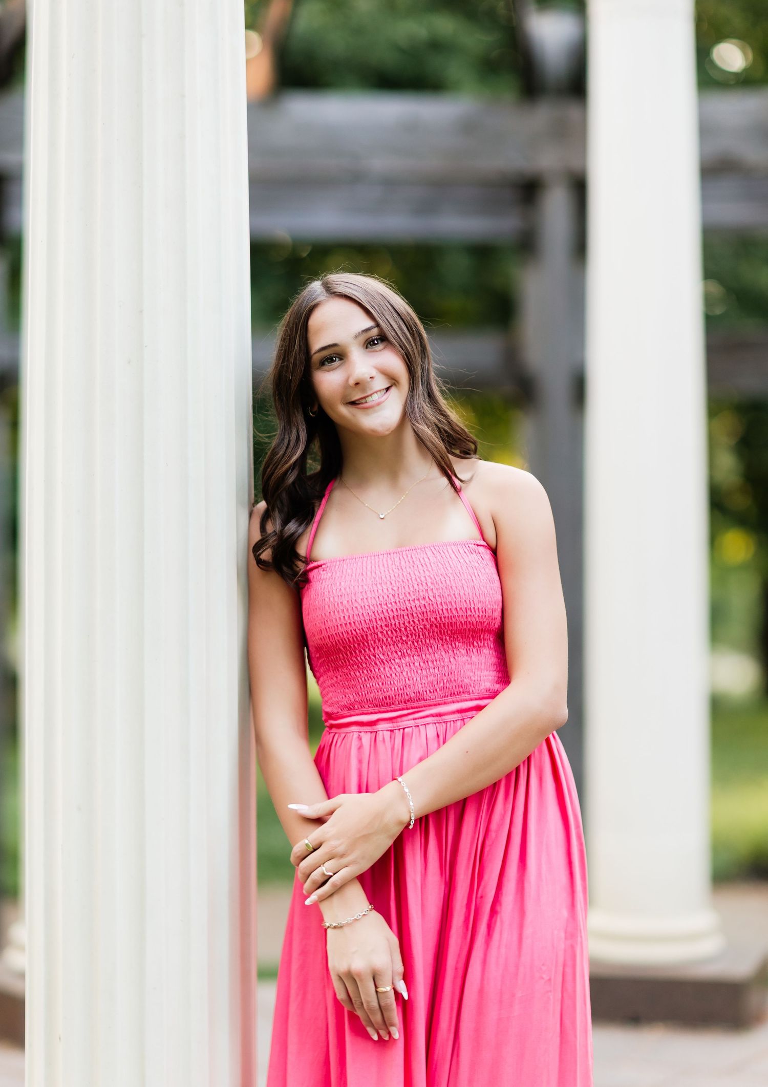 Young woman in pink smocked dress smiling while leaning against white outdoor column.