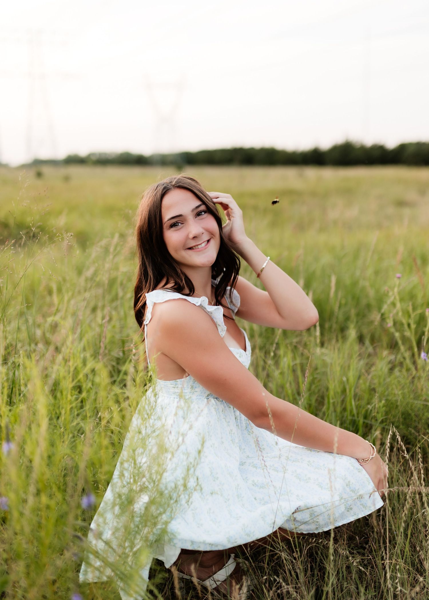Smiling brunette teen in white floral dress sitting in a tall green meadow field outdoors.