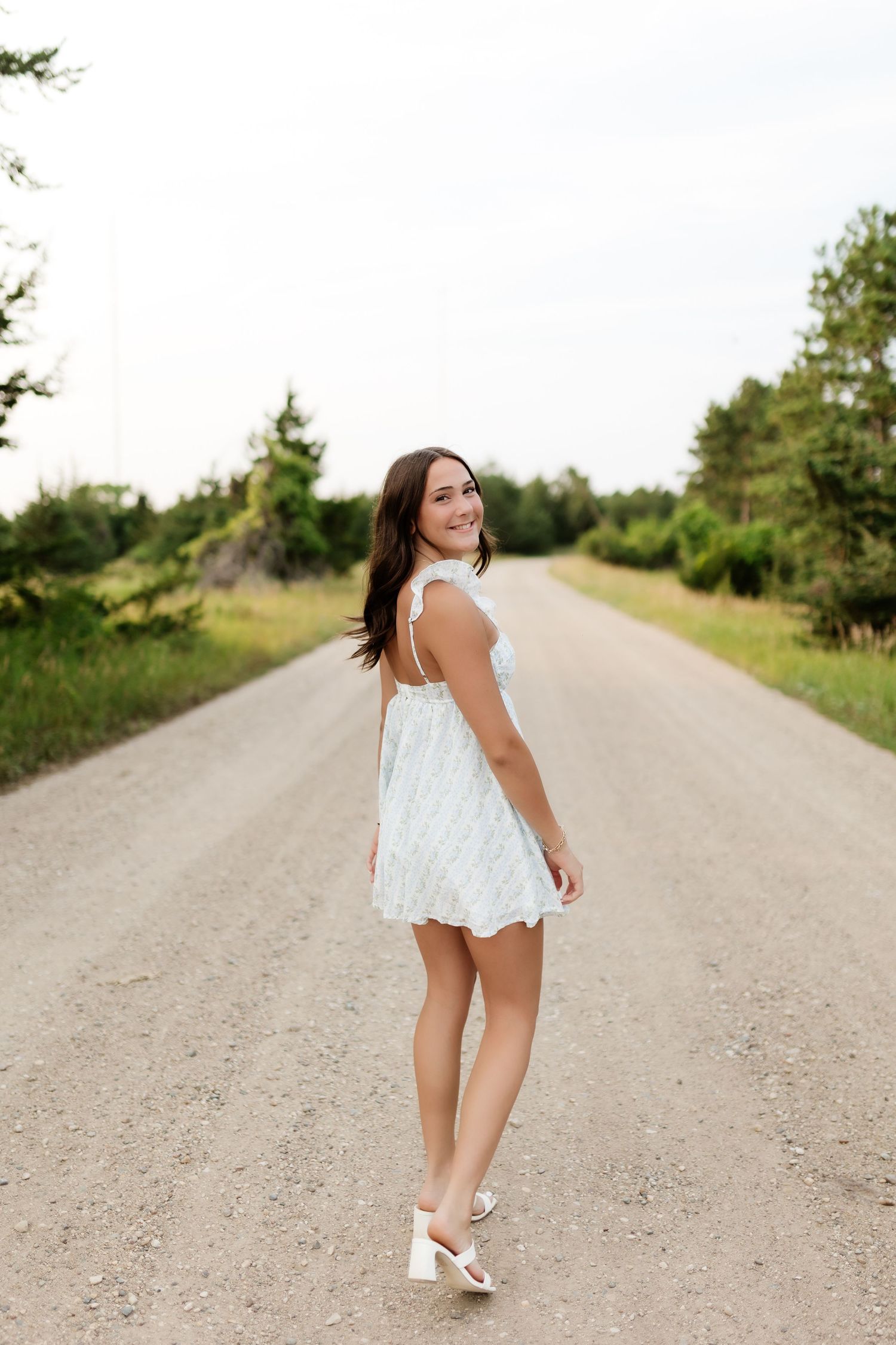 Smiling young woman in a white mini dress walks along a scenic tree-lined gravel road on a summer day.