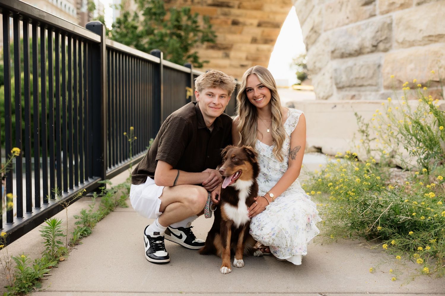Couple crouching with their Australian Shepherd dog near a stone wall and black iron fence outdoors.