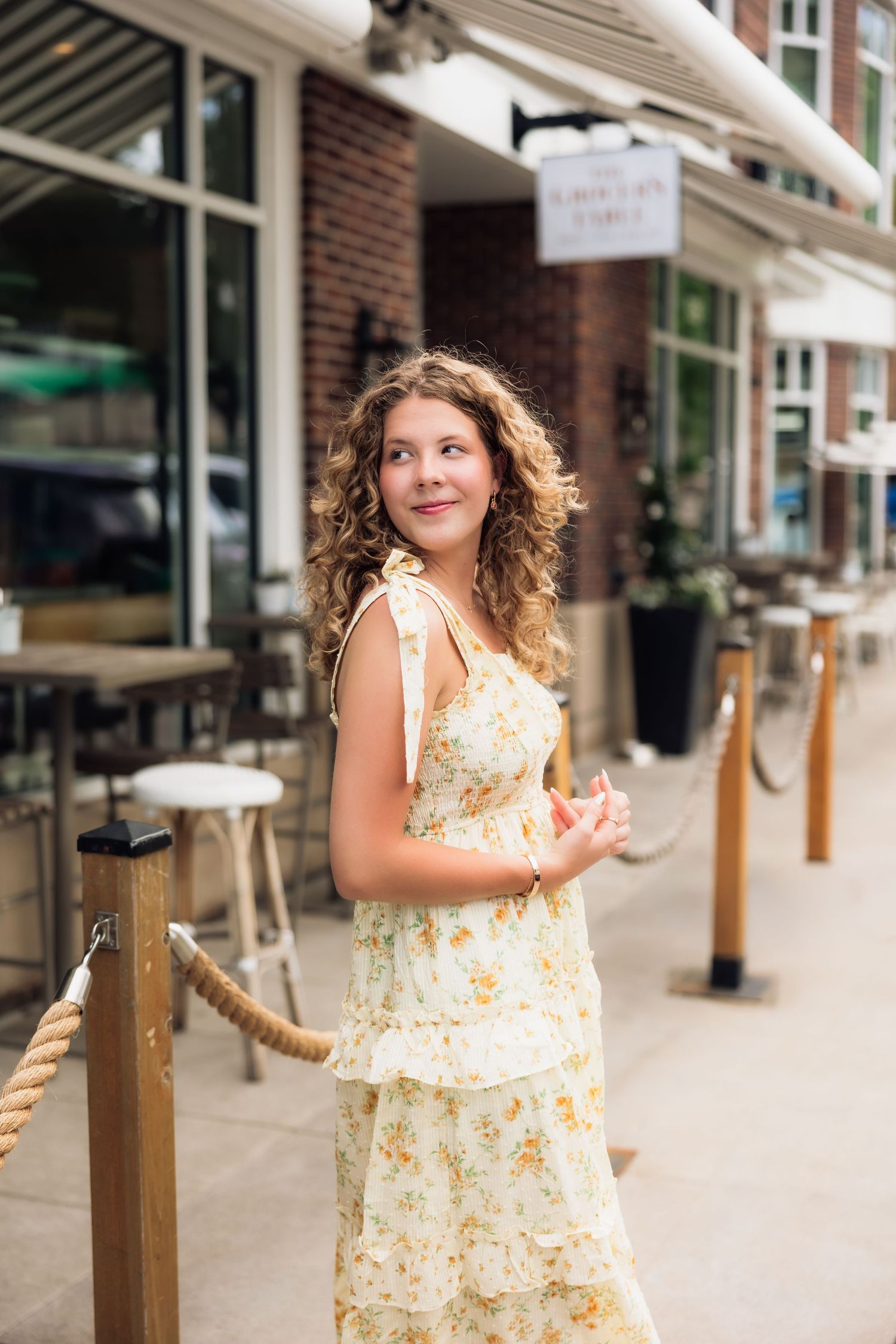 Curly-haired young woman in a yellow floral midi dress posing on a sidewalk outside a restaurant.