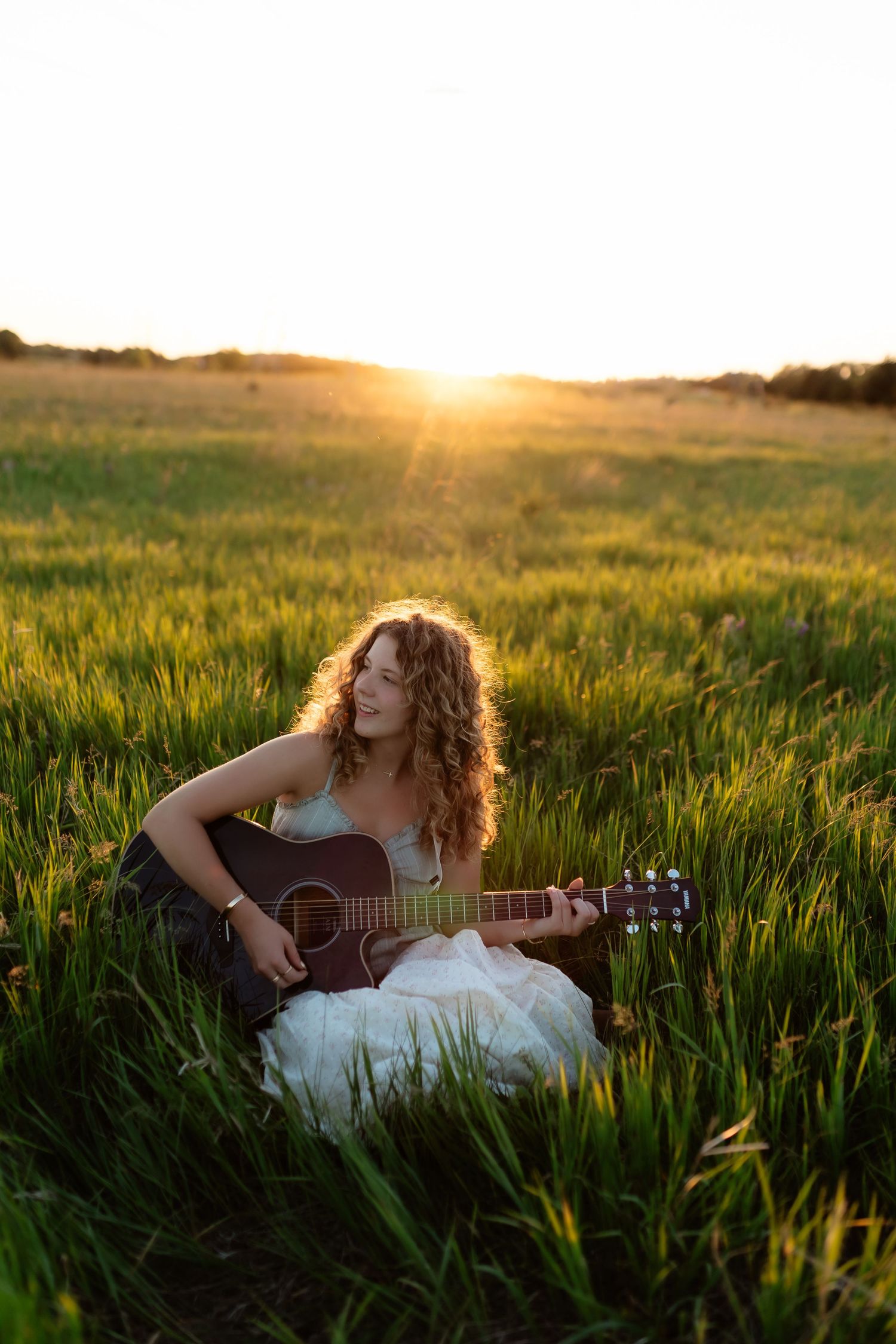 Young woman with curly hair playing acoustic guitar while sitting in green field at golden sunset.
