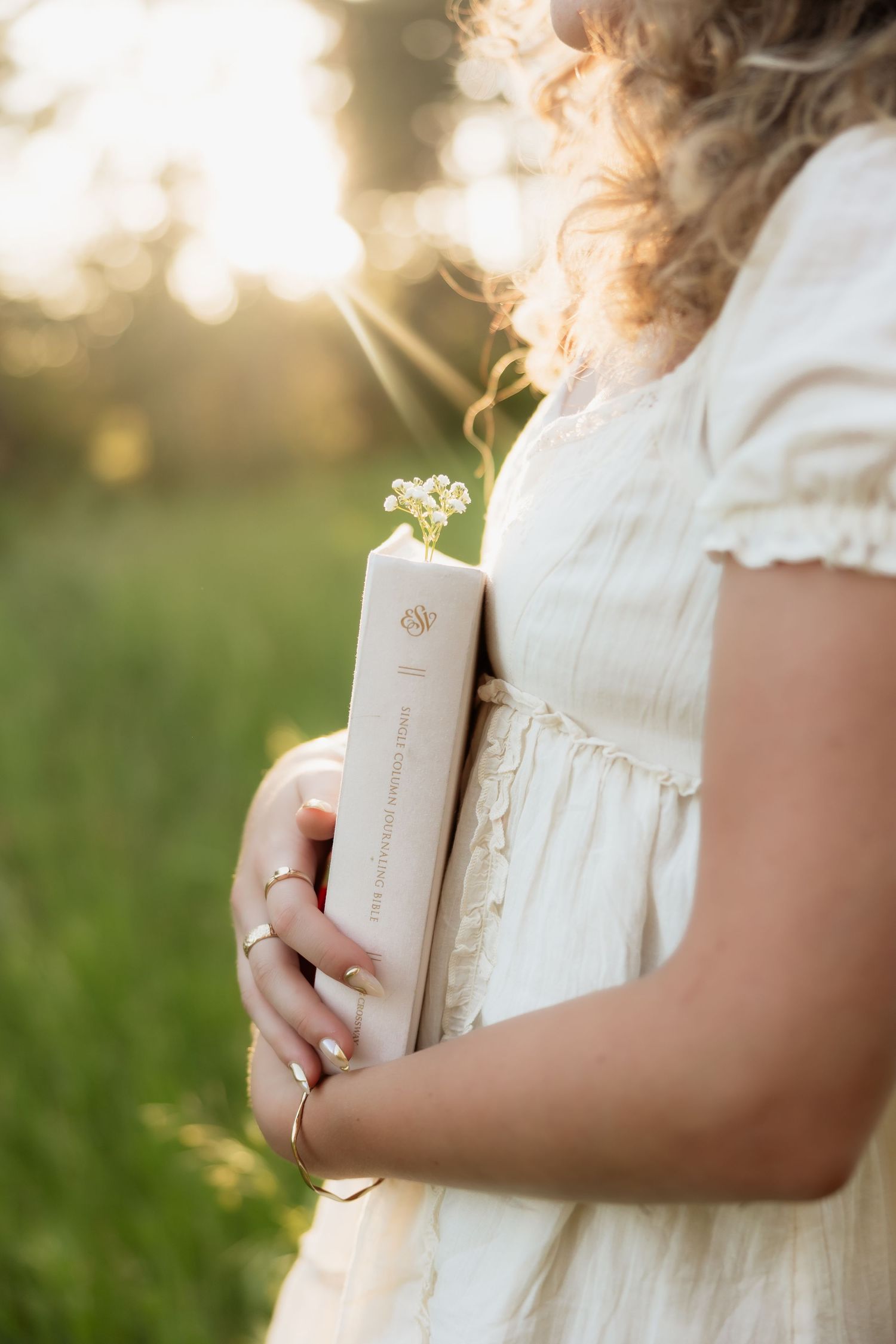 Close-up of a woman in cream dress holding a book with a small wildflower in golden sunset light.