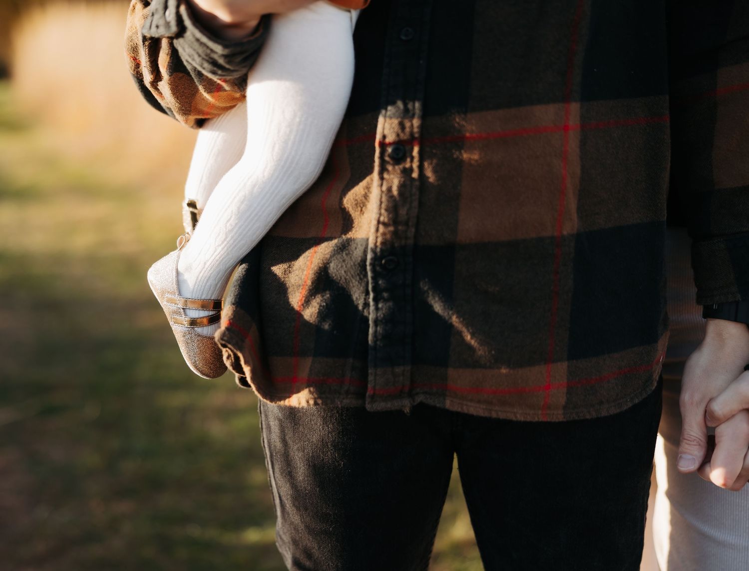 Father in plaid flannel shirt holding a baby dressed in white against a warm outdoor autumn background.