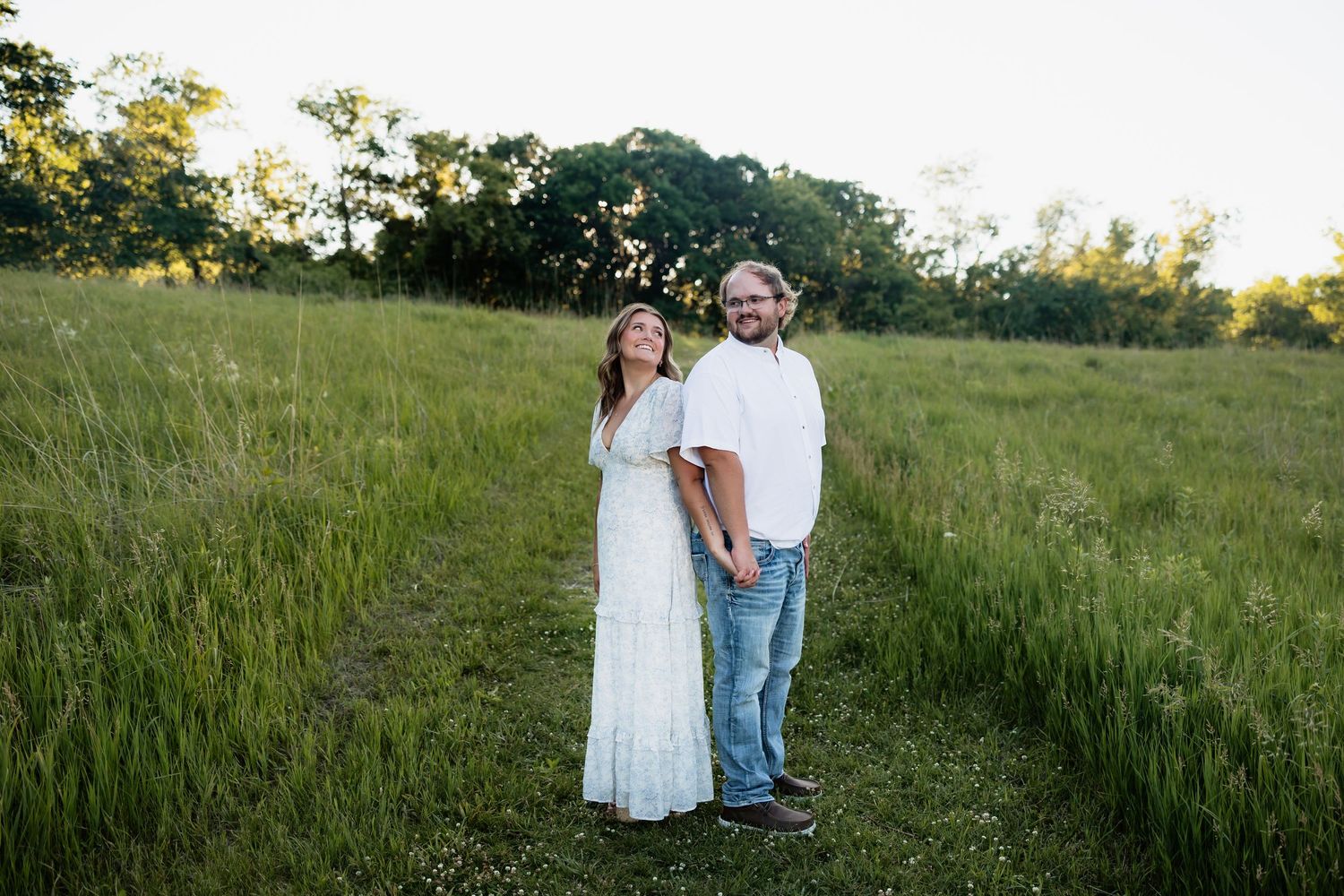 Couple holding hands in a green meadow at golden hour, woman in white dress, man in jeans and white shirt.