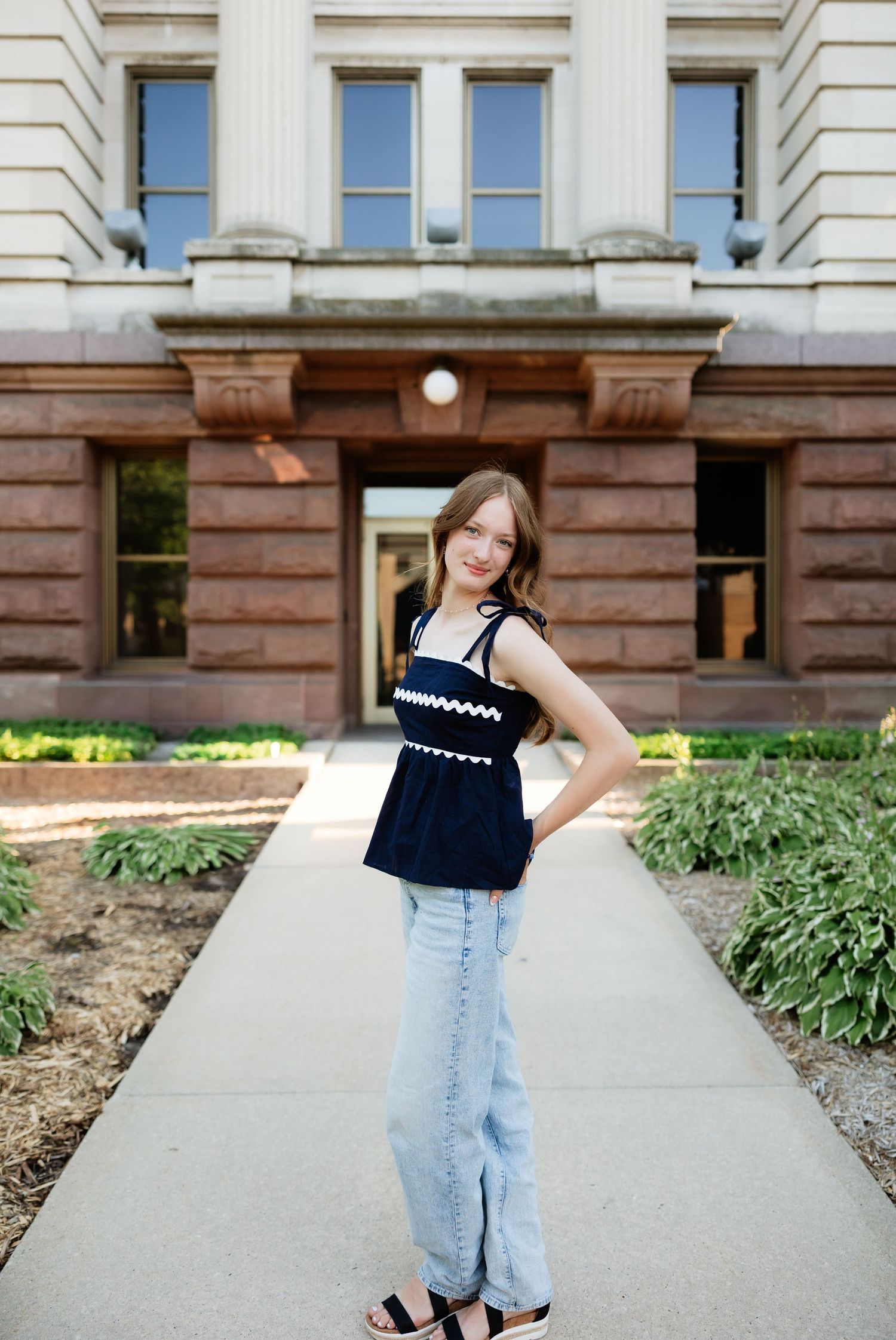 Young woman in navy top and light jeans poses on sidewalk outside historic stone building.