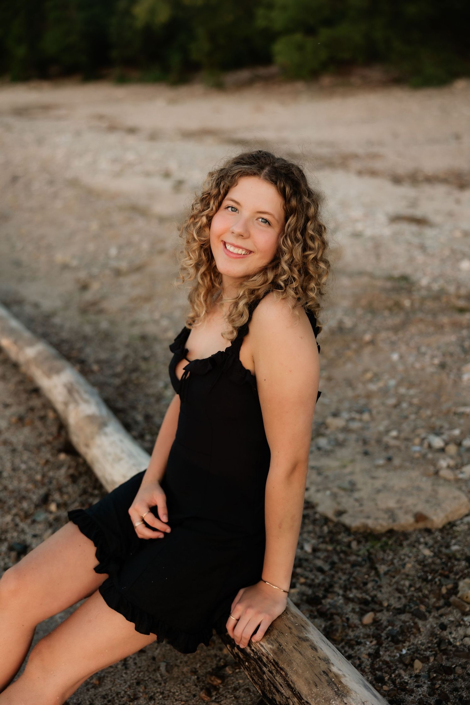 Young woman with curly hair in black dress sitting on driftwood at a rocky beach, smiling outdoors.