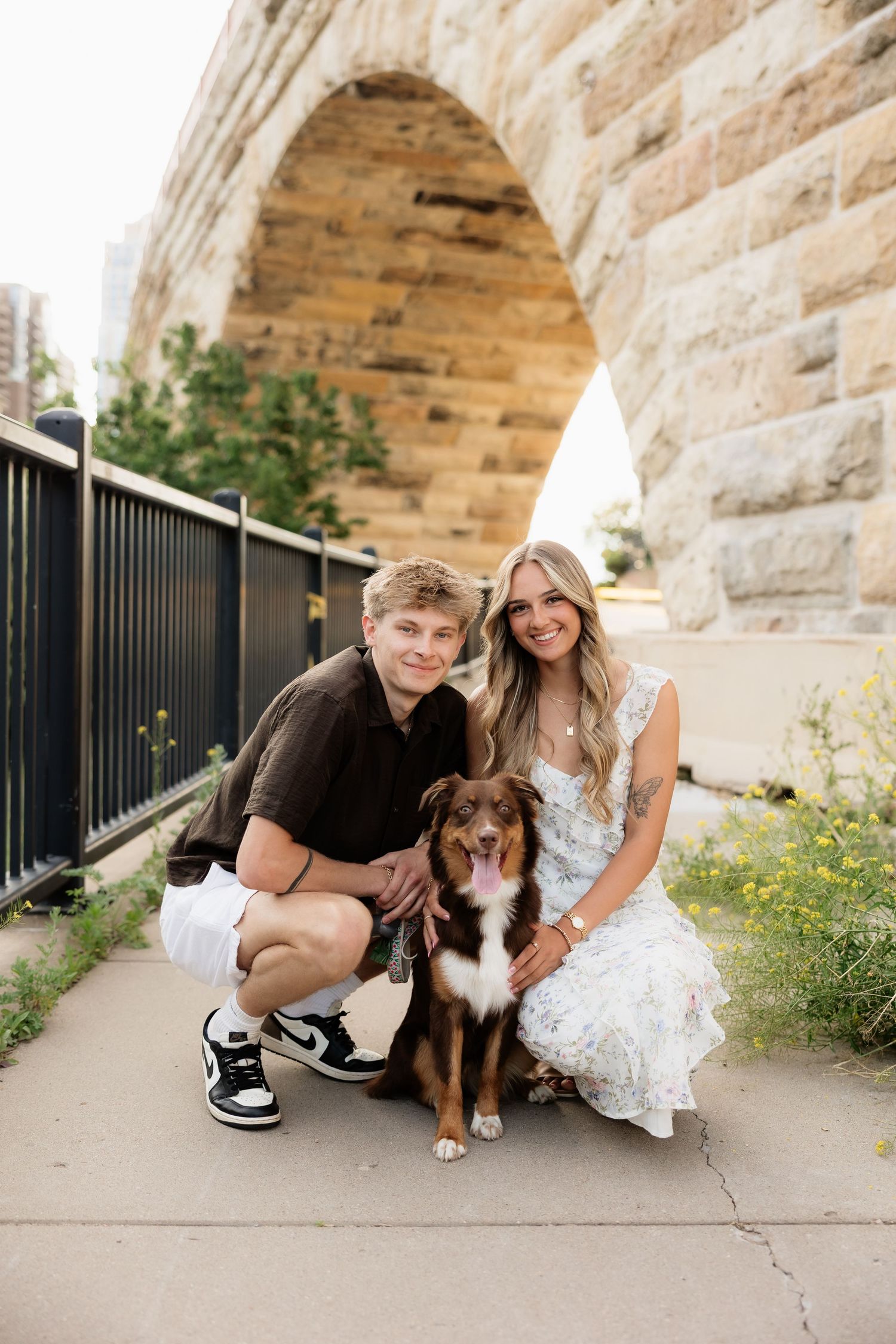 Couple crouching with their Australian Shepherd dog in front of a stone arch bridge on a sunny day.