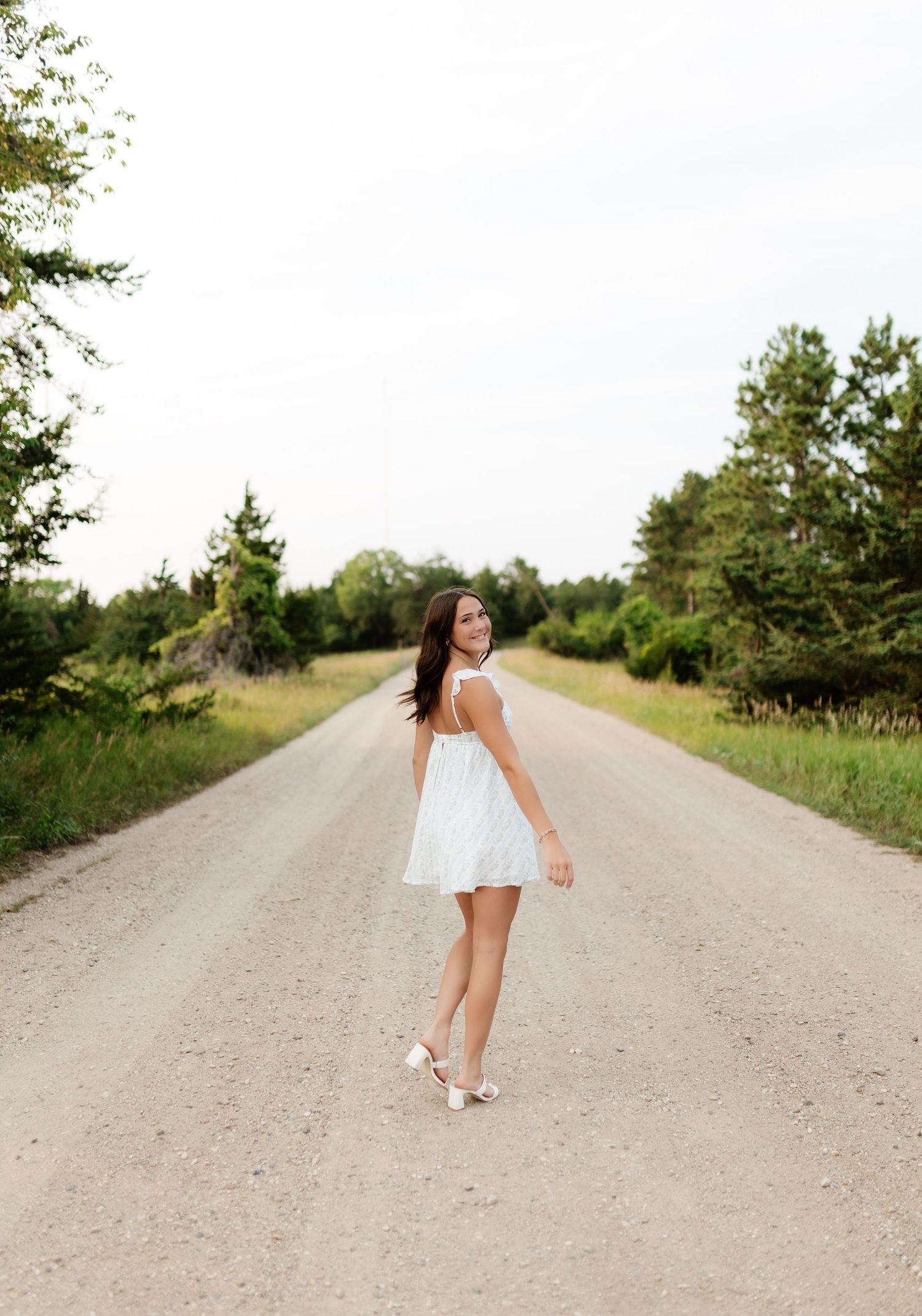 Young brunette woman in white mini dress walking on a scenic tree-lined gravel road.