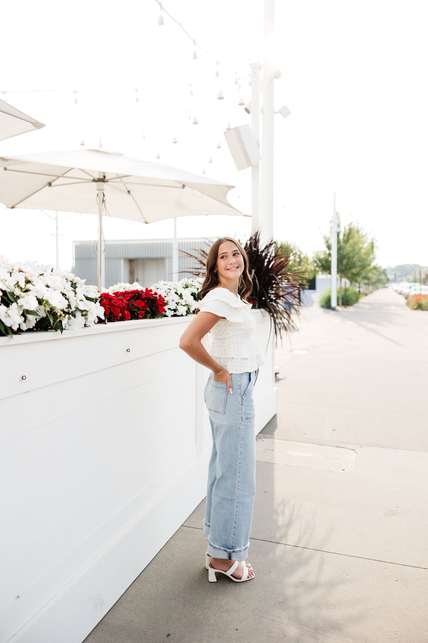 Smiling young woman in white ruffled top and jeans posing by outdoor floral planters on a sunny sidewalk.