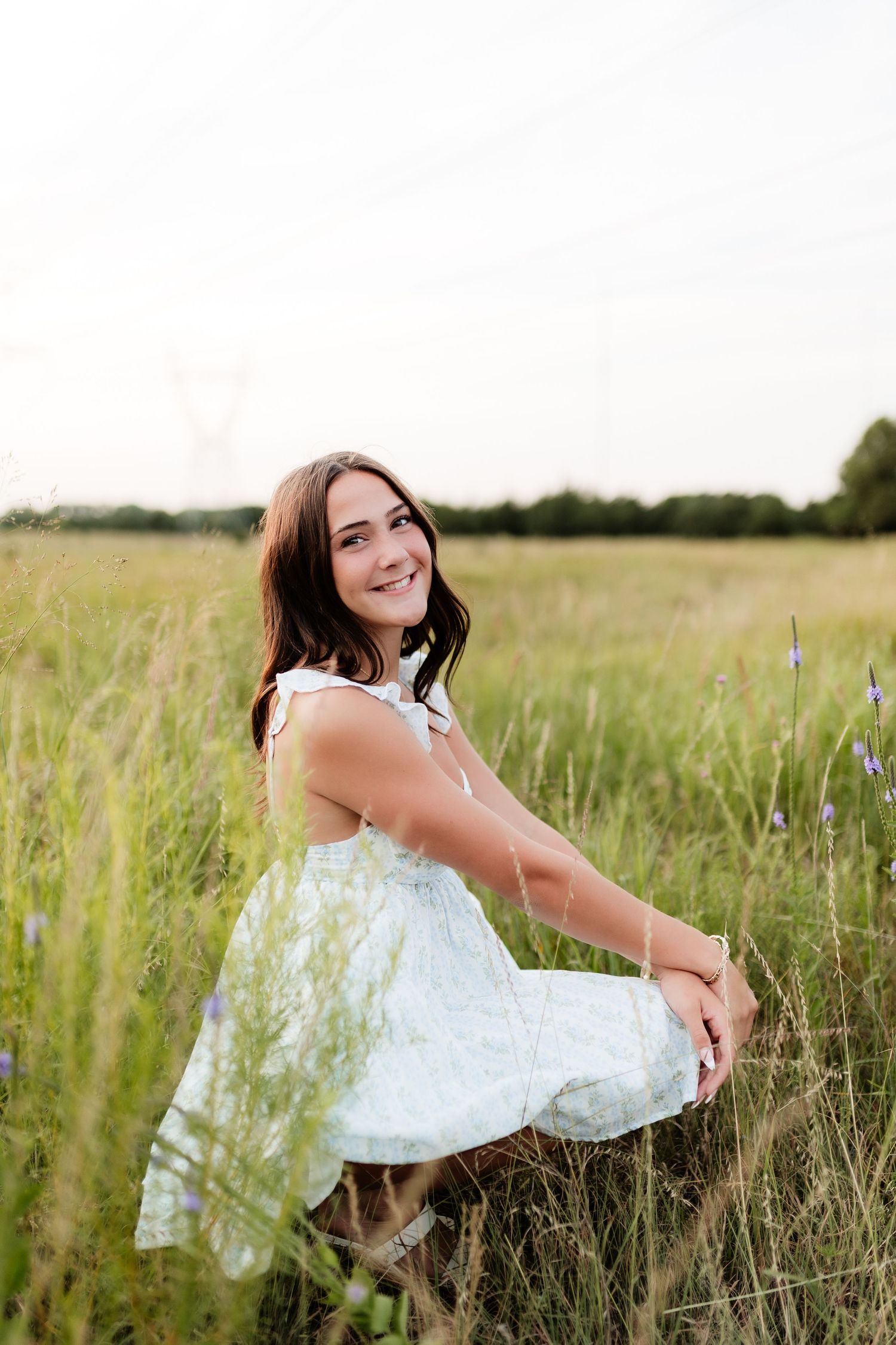 Smiling young woman in a white floral dress sits in a tall green meadow on a bright summer day.