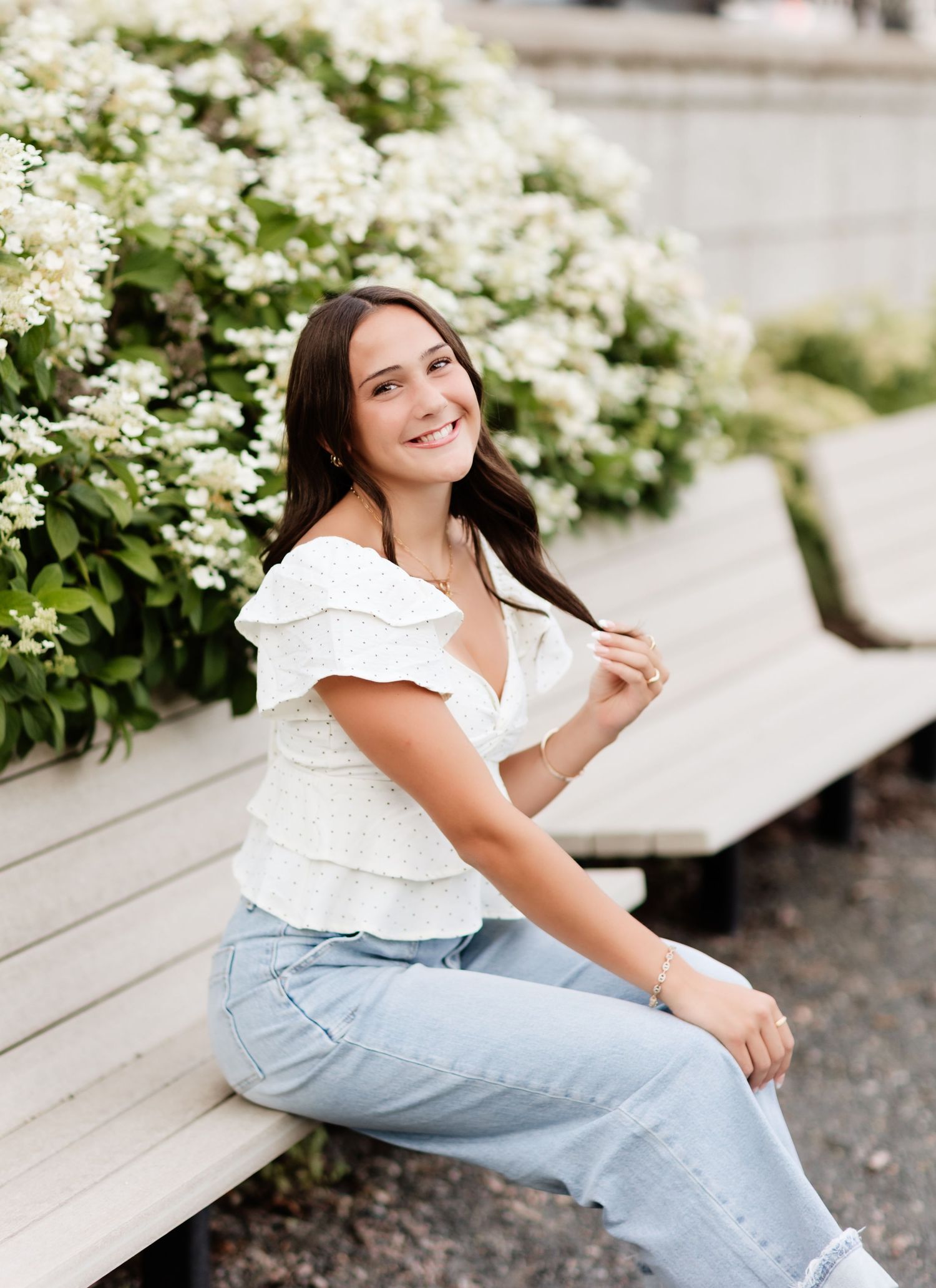 Brunette woman in white ruffled top and light jeans sitting on bench beside white flowering bush.