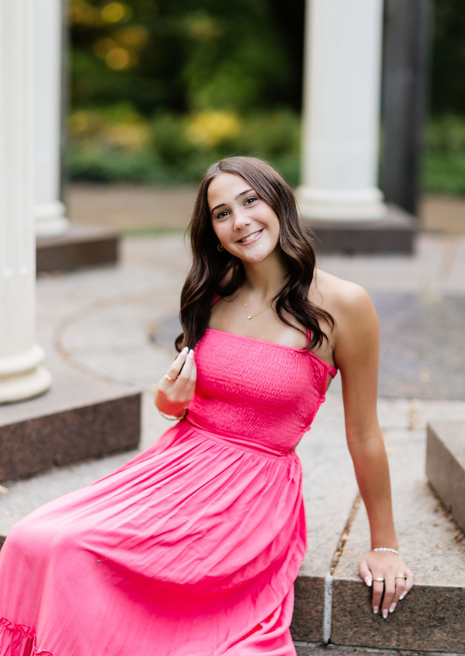 Young woman in strapless pink dress seated on steps near white columns, smiling in outdoor setting.