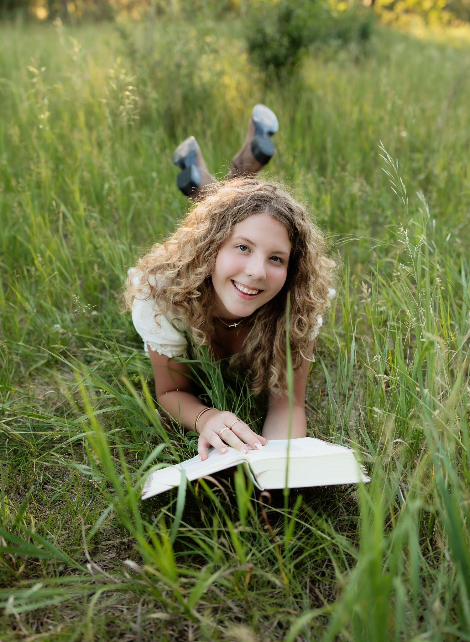 Smiling curly-haired girl lying in green grass reading an open book outdoors.