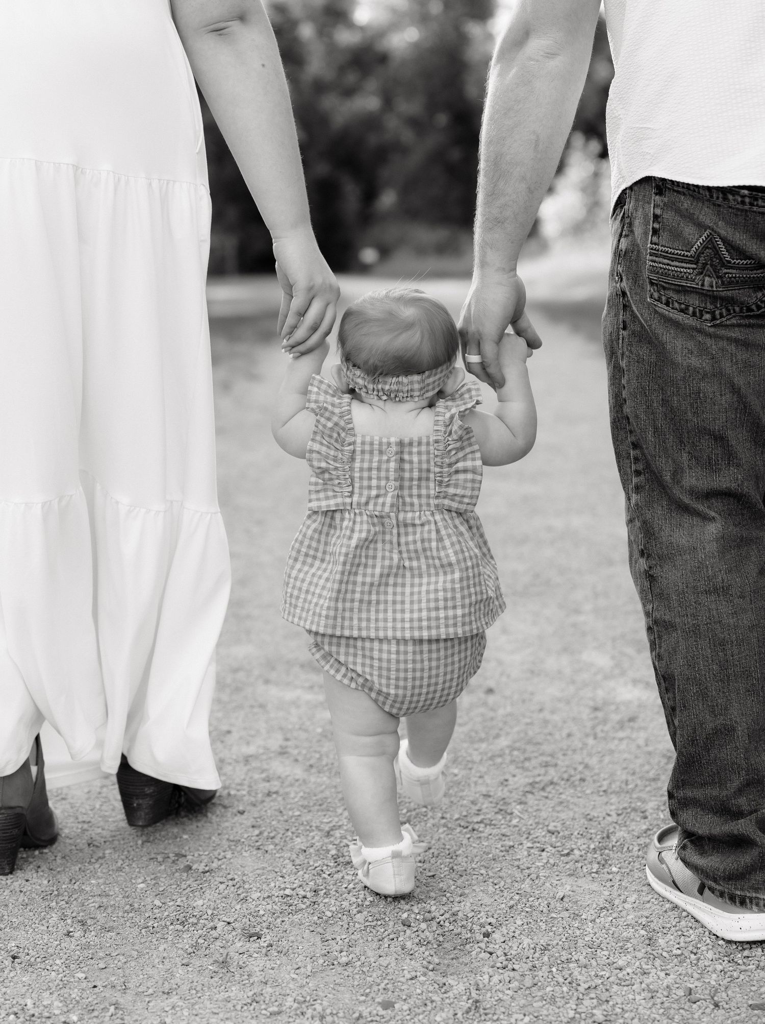 Black and white photo of a baby taking first steps while holding parents hands on a gravel path outdoors.