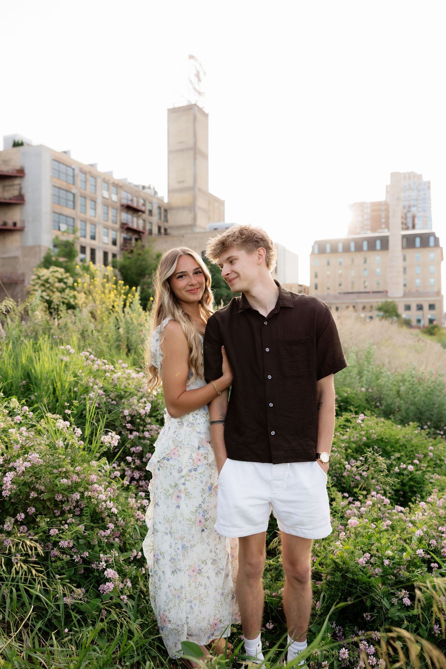 A smiling couple standing together in a lush urban garden, woman in floral maxi dress, man in black shirt and white shorts.