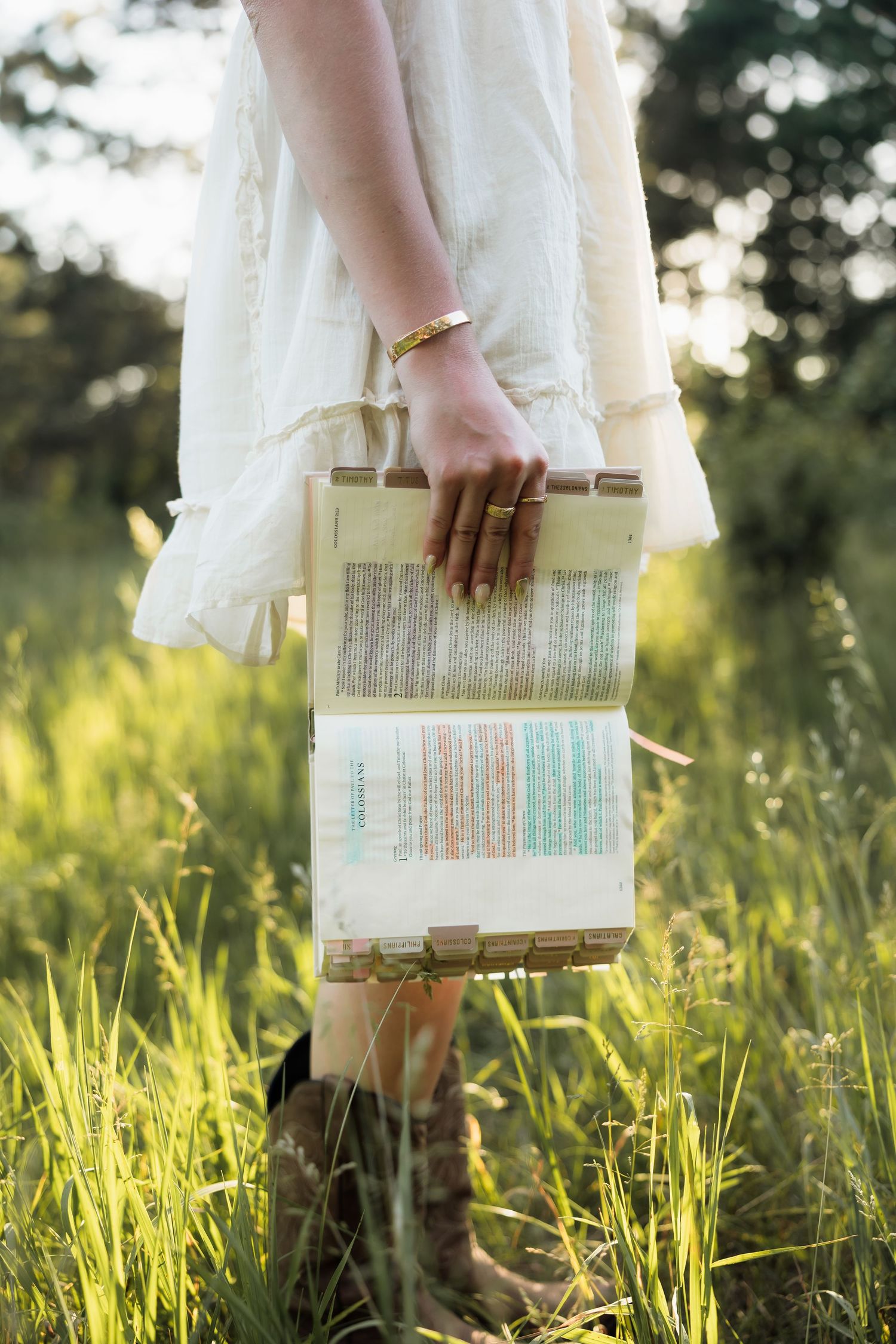 Woman in white dress and cowboy boots holding a decorative woven textile artwork in a sunny green meadow.