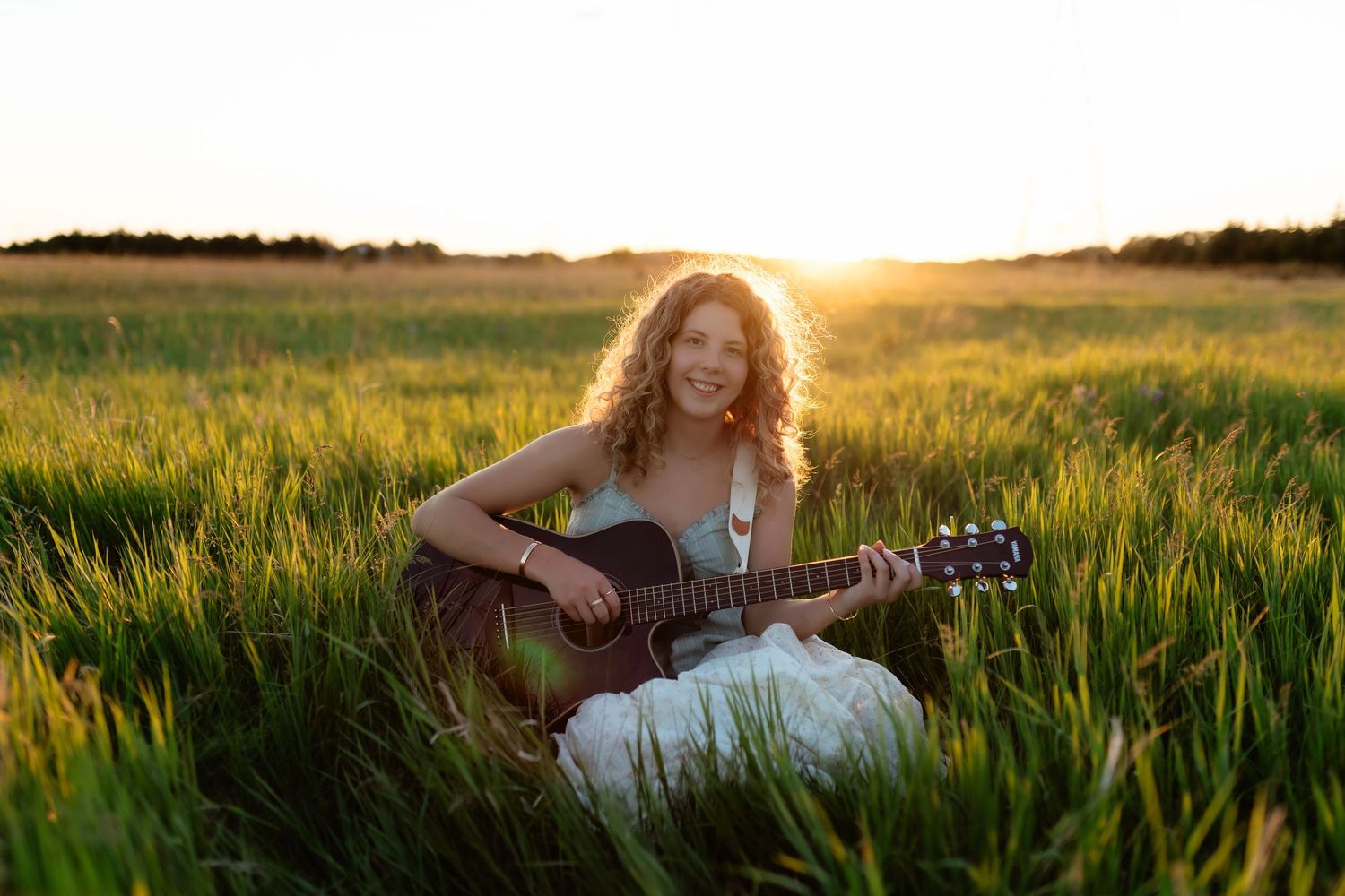 Smiling young woman playing acoustic guitar while sitting in lush green field at golden sunset.