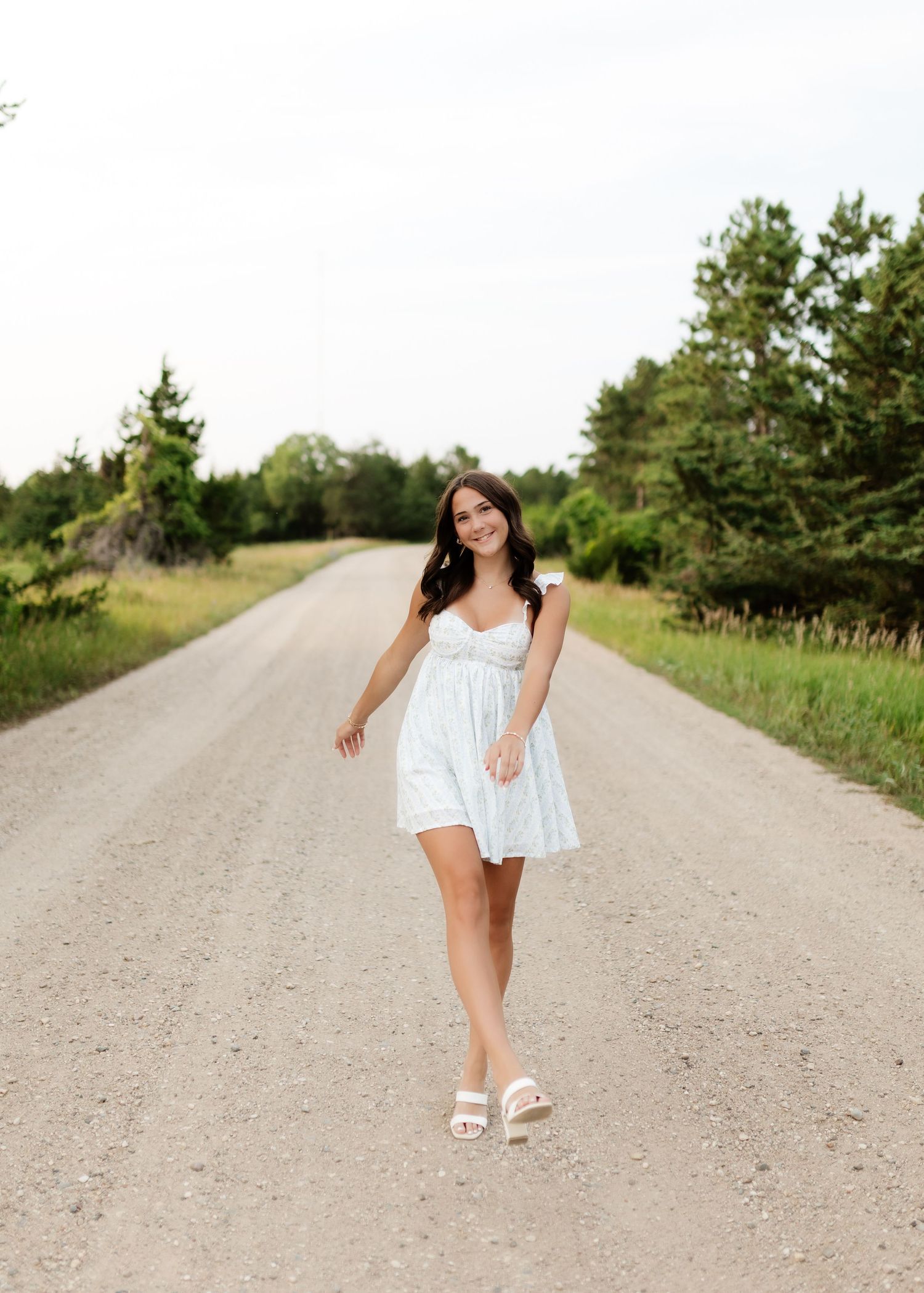 Smiling brunette woman in white mini dress walking on rural gravel road lined with trees.