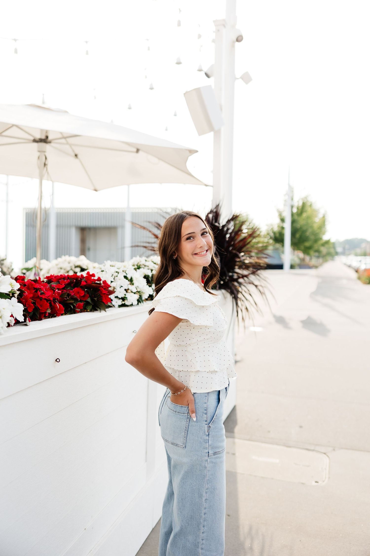 Young woman in white top and denim skirt posing beside white planter with red and white flowers.